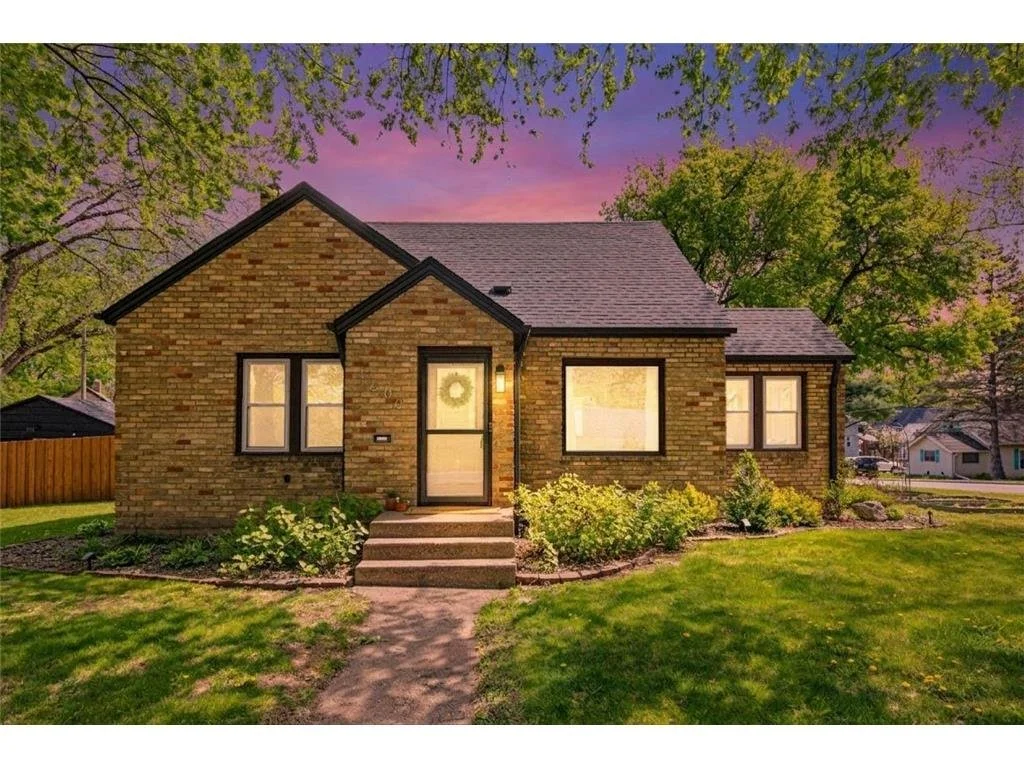 A cozy brick house with a front porch, surrounded by well-maintained lawn and landscape, under a colorful evening sky with trees nearby.