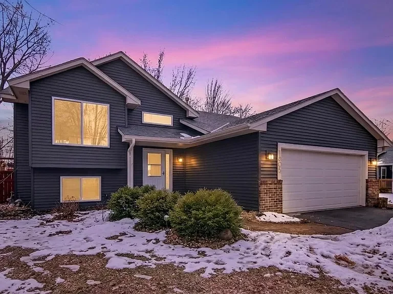 A two-story blue house with a two-car garage, illuminated exterior lights, and snow on the front yard during sunset.