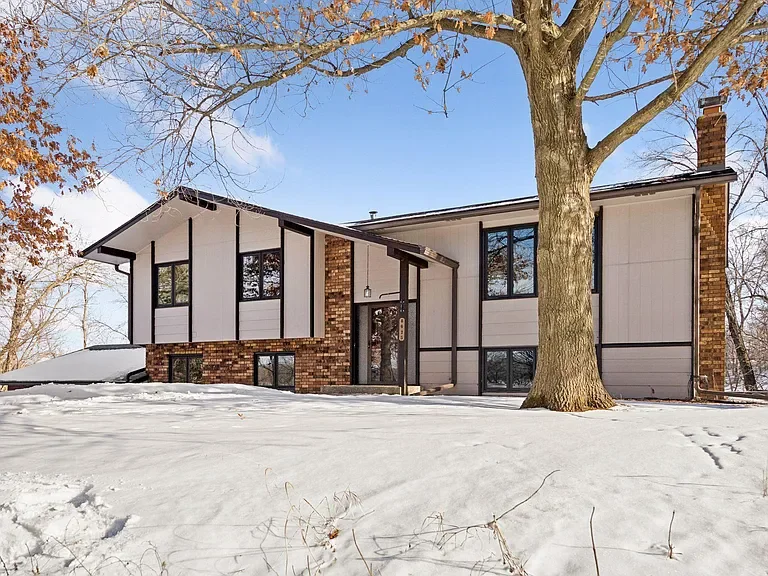 A modern two-story house with large windows, brick accents, and a sloped roof, surrounded by snow and a large leafy tree in the foreground.