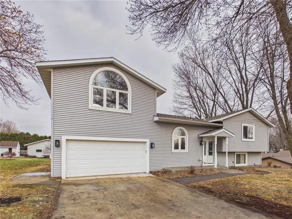 A two-story suburban house with beige siding, a white garage door, and multiple windows, including a large arched window on the upper floor. The front yard has a small porch with columns, and the driveway is made of concrete. Bare trees surround the house, indicating a late fall or winter season.