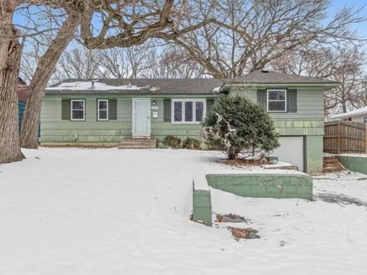 A green single-family house with snow-covered front yard, steps leading to the front door, and a large bush near the house. Trees with bare branches are visible in the background.