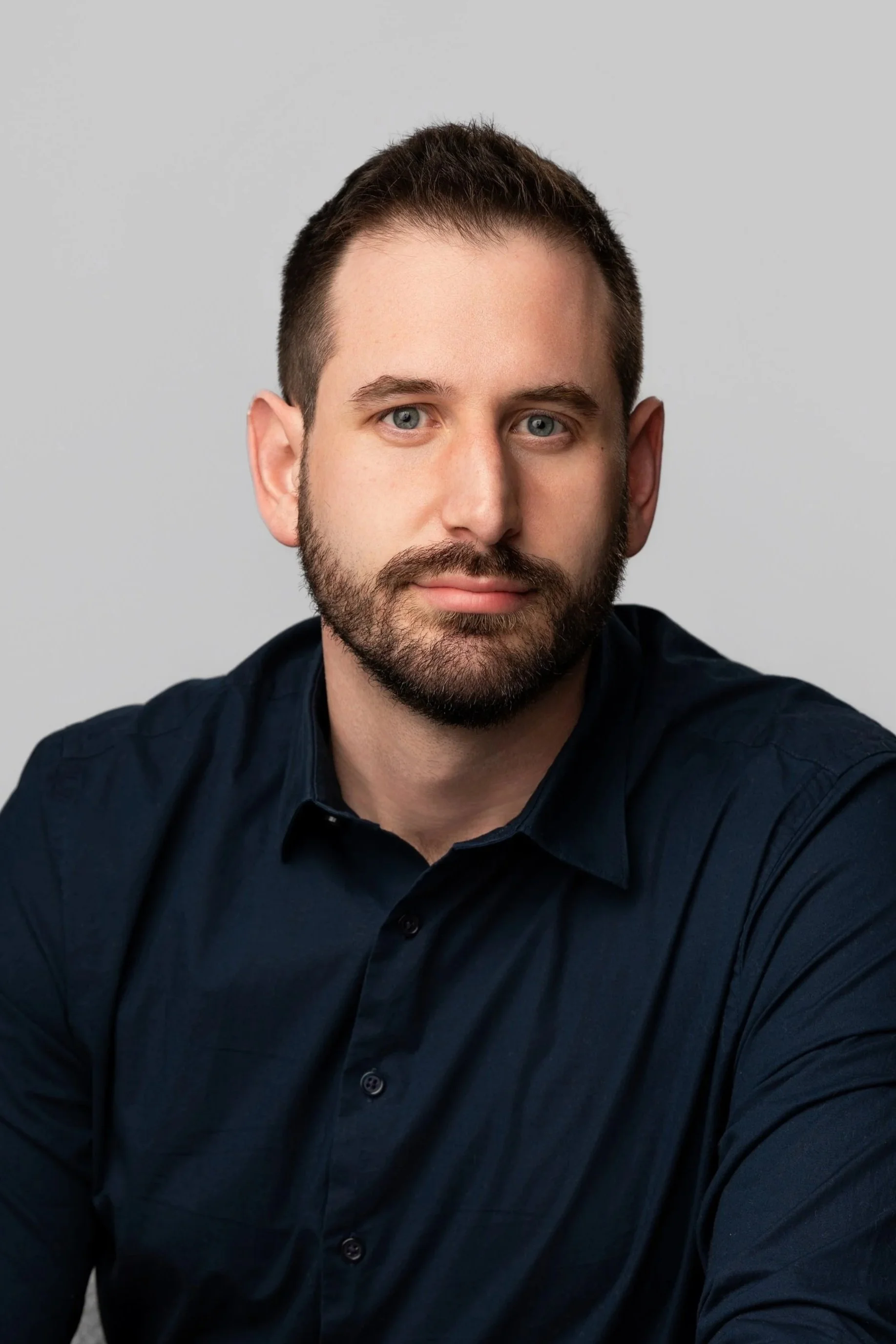 Close-up portrait of a man with short, dark hair and a beard, wearing a dark blue shirt, against a plain gray background.