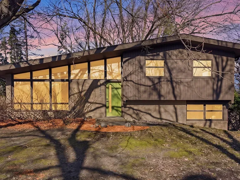 Modern house with black exterior, large windows, and a bright green front door, surrounded by trees with shadows cast on the ground during sunset.