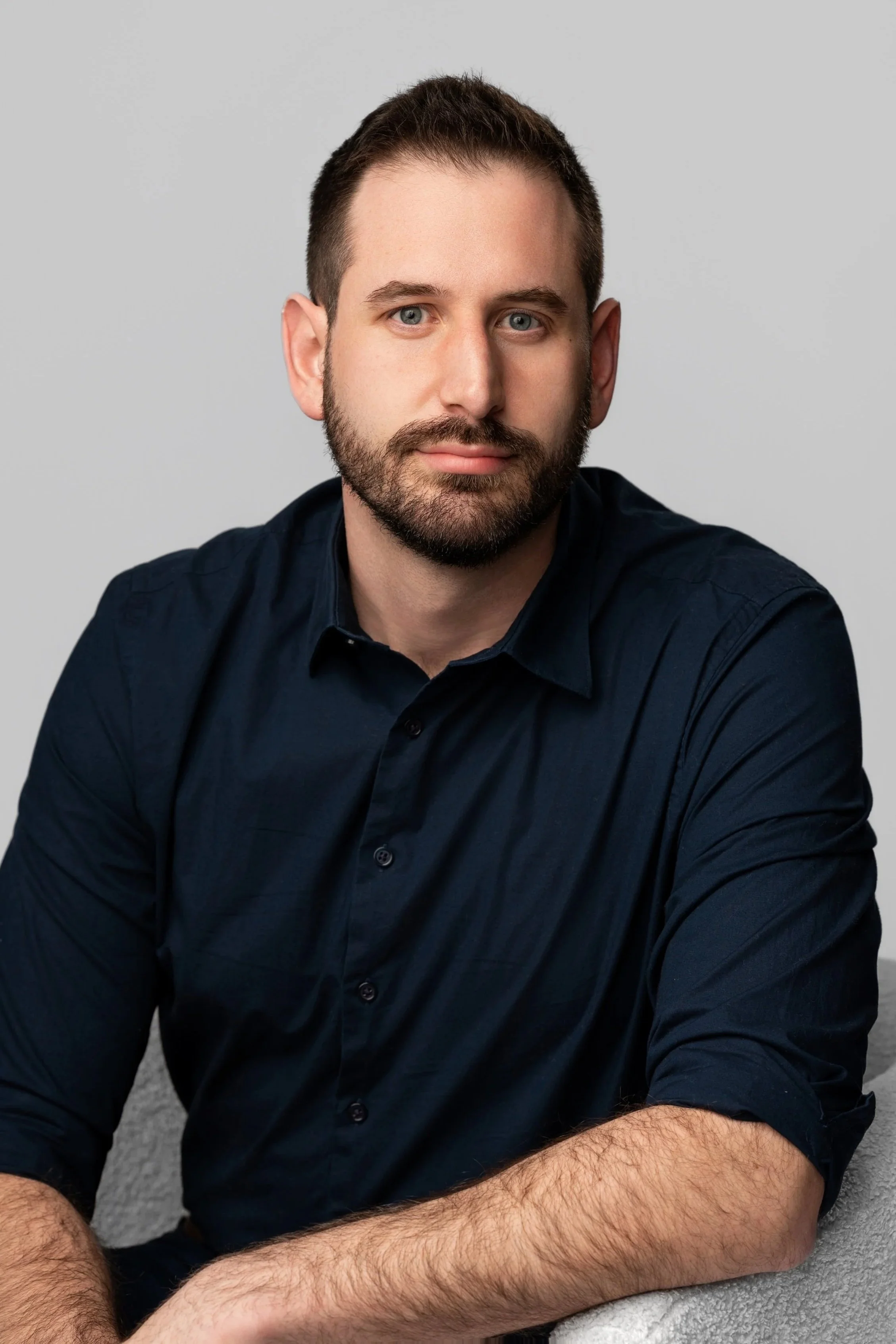 A man with short brown hair and a beard sitting against a light grey background, wearing a dark blue shirt.