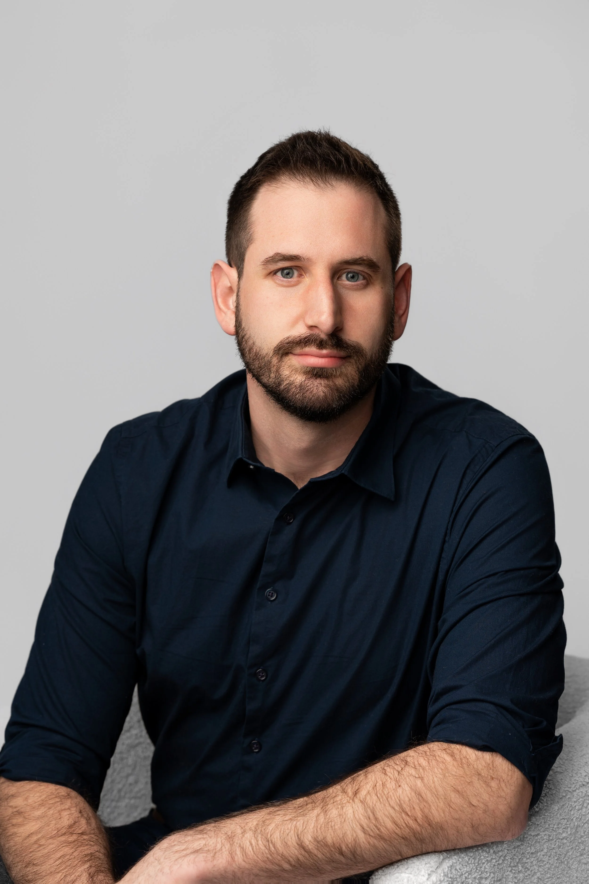A man with short brown hair, a beard, and blue eyes sitting on a gray carpeted floor in front of a plain gray wall, wearing a black button-up shirt with the sleeves rolled up.