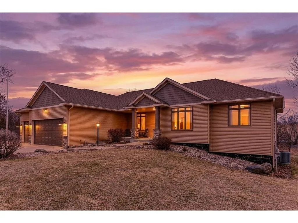 A modern single-story house with a garage, light exterior, lit windows, and a front porch illuminated at sunset.