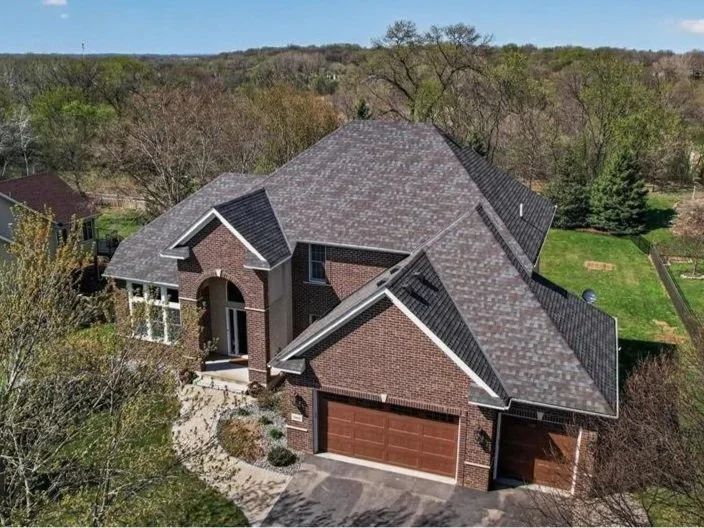 Aerial view of a large brick house with a gray shingled roof, front porch, attached garage, surrounded by trees and green yard.