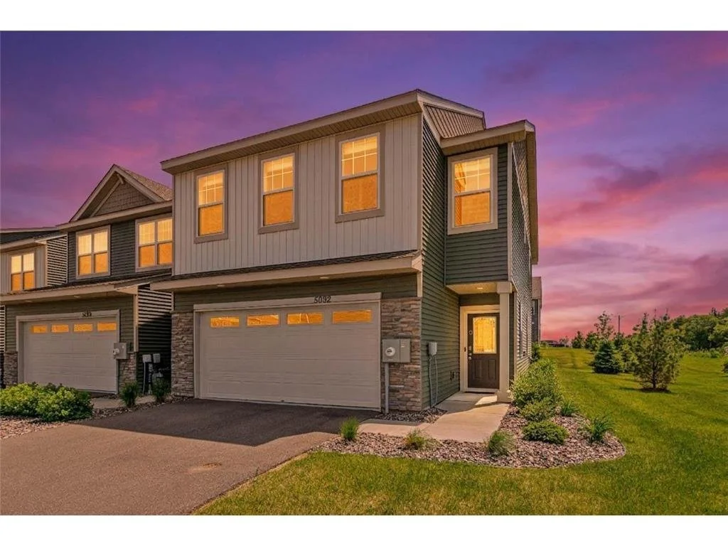 A modern two-story house with a garage, illuminated from inside, during sunset. The house has gray siding with stone accents, a black door, and a well-maintained green lawn with small shrubs.