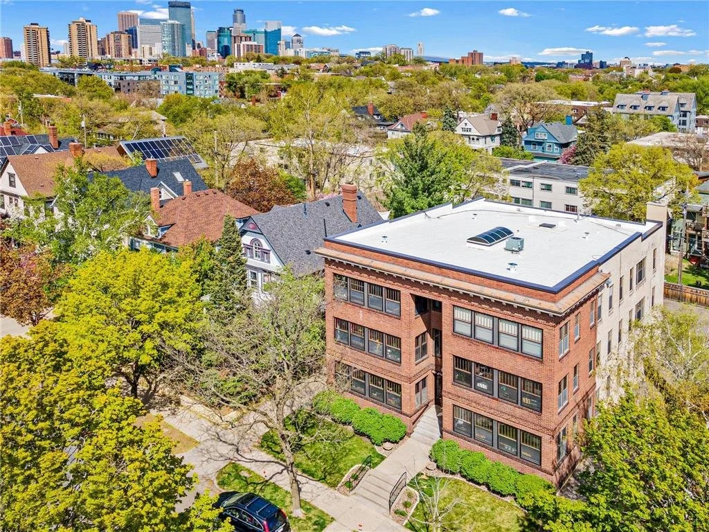 An aerial view of a multi-story brick building surrounded by trees and a sidewalk in a residential area with single-family houses and a distant city skyline under a partly cloudy sky.