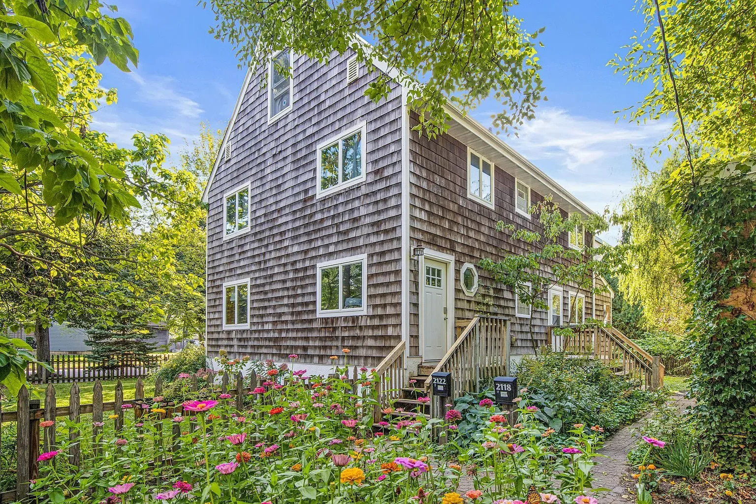 A wooden house with a sloped roof and multiple windows, surrounded by a garden filled with pink, yellow, and purple flowers, and green trees, under a blue sky with some clouds.