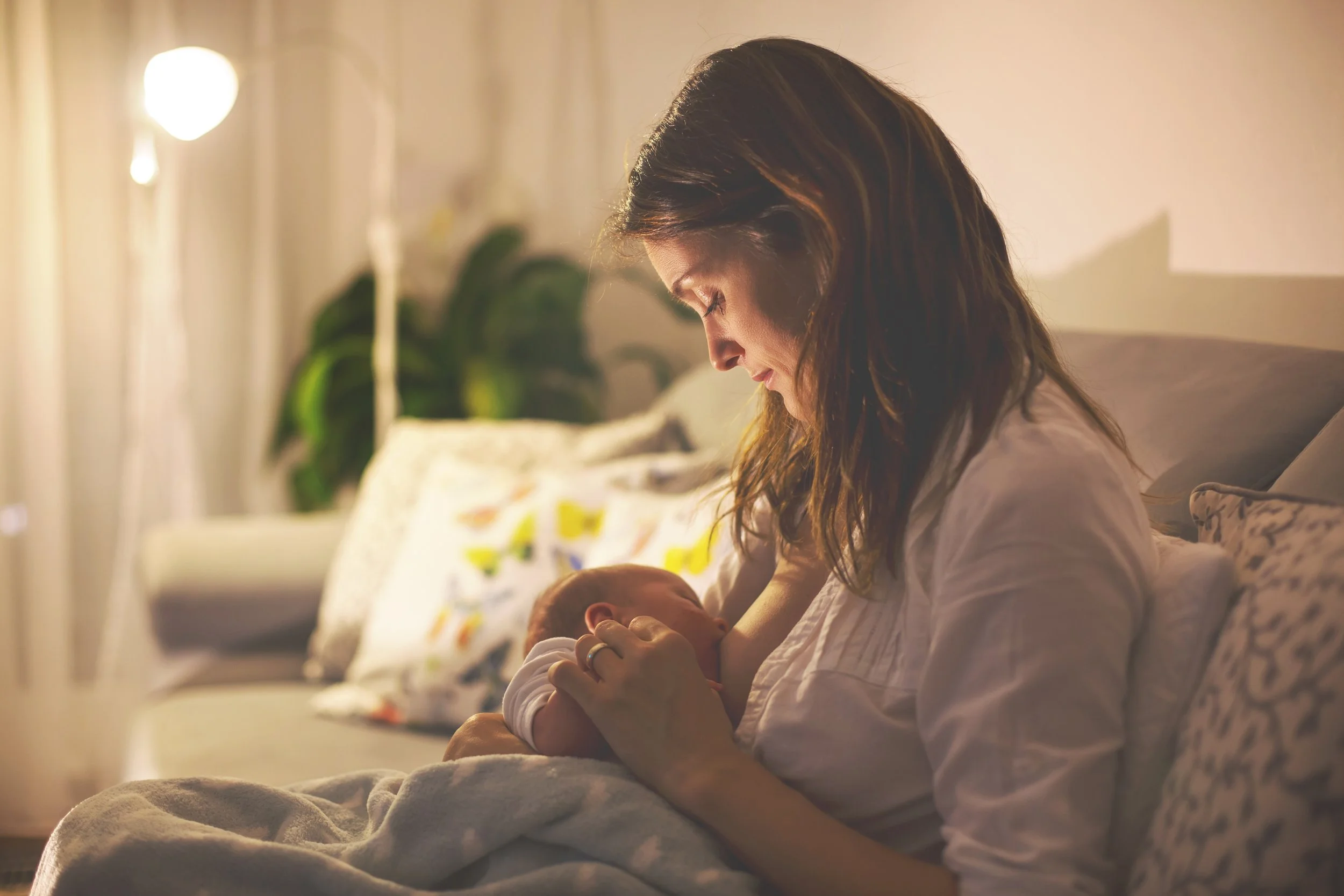A woman sitting on a bed, gently holding and looking at a newborn baby in her arms, in a warmly lit room with a houseplant and decorative pillows in the background.