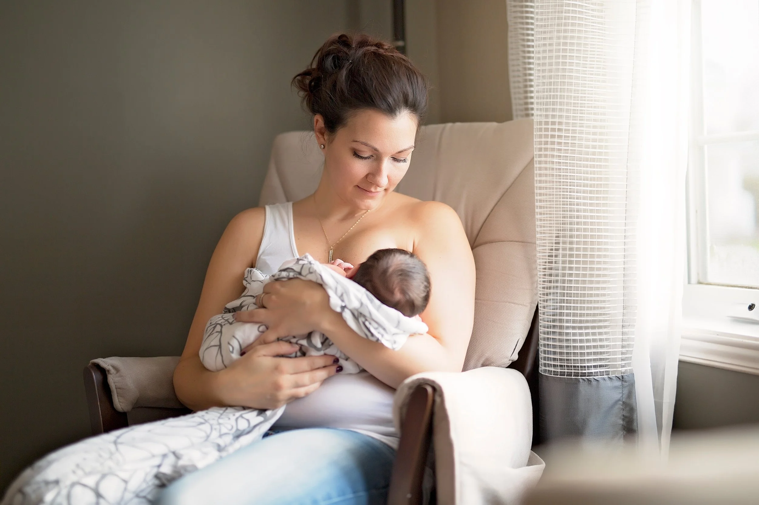 A woman with dark hair in a bun sitting on a beige armchair by a window, breastfeeding a baby wrapped in a patterned blanket.