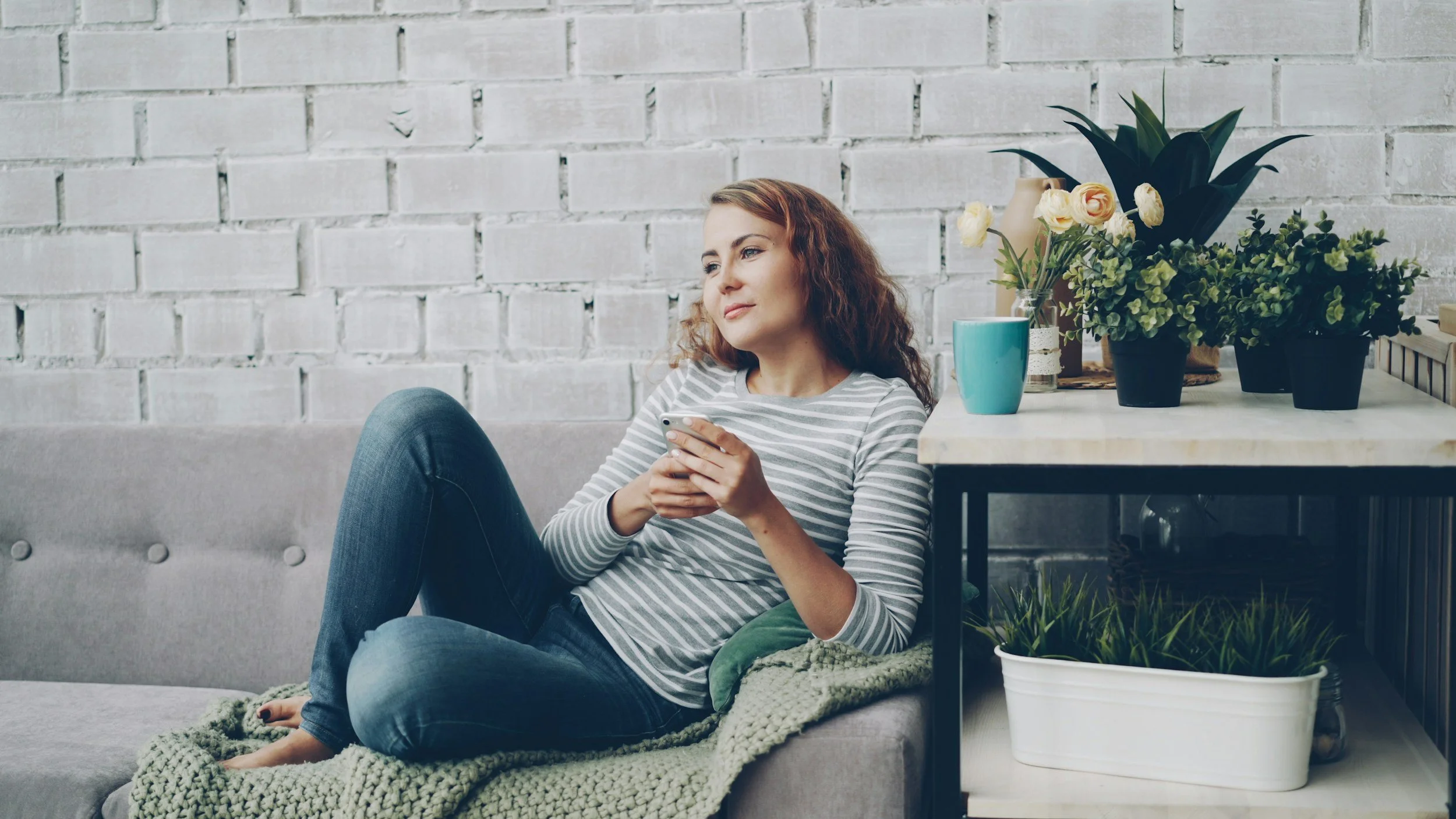 Woman sitting on a gray couch, looking at her smartphone, with plants and flowers on a side table beside her, against a white brick wall.