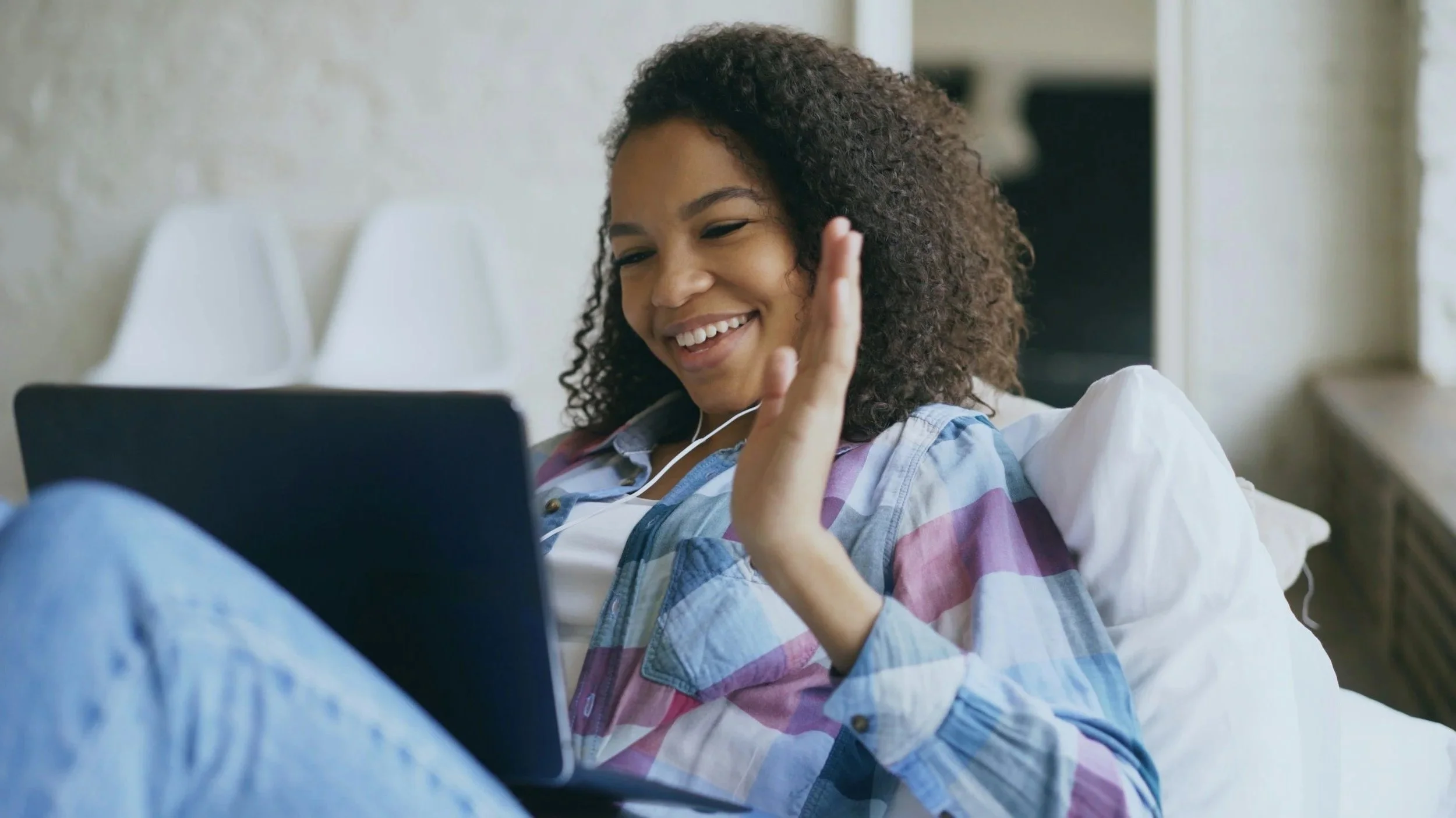 A young woman with curly hair smiling and waving while looking at her tablet. She is sitting on a white sofa in a bright room, wearing a colorful striped shirt.