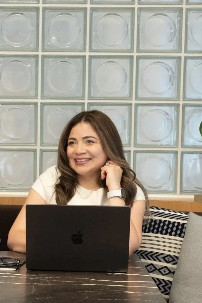 A woman with long brown hair smiling and sitting at a table with a black MacBook in front of her. She is wearing a white top and a smartwatch. The background features glass blocks.