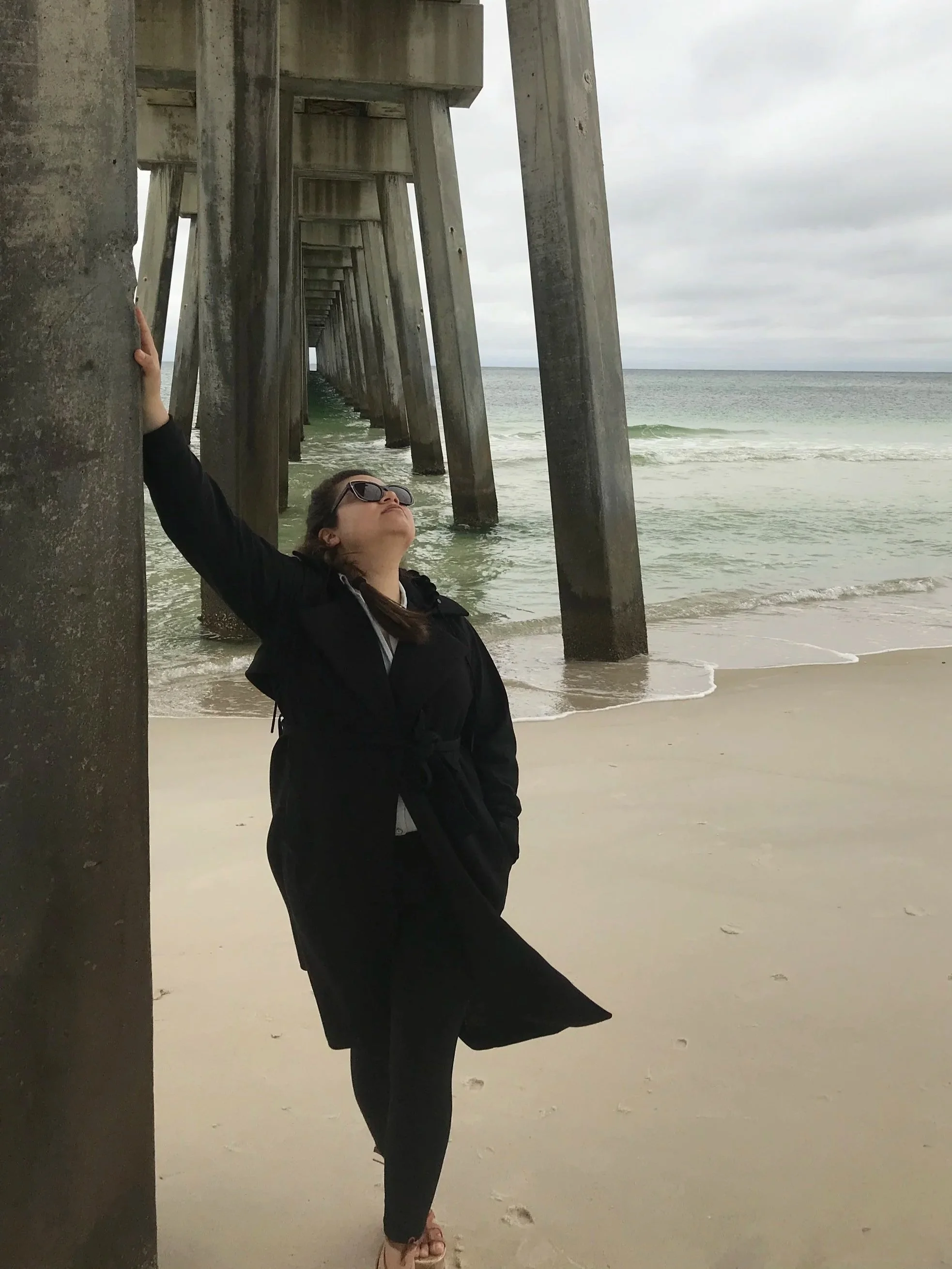 A woman wearing sunglasses and a black jacket standing under a pier at the beach during overcast weather, with the ocean in the background.