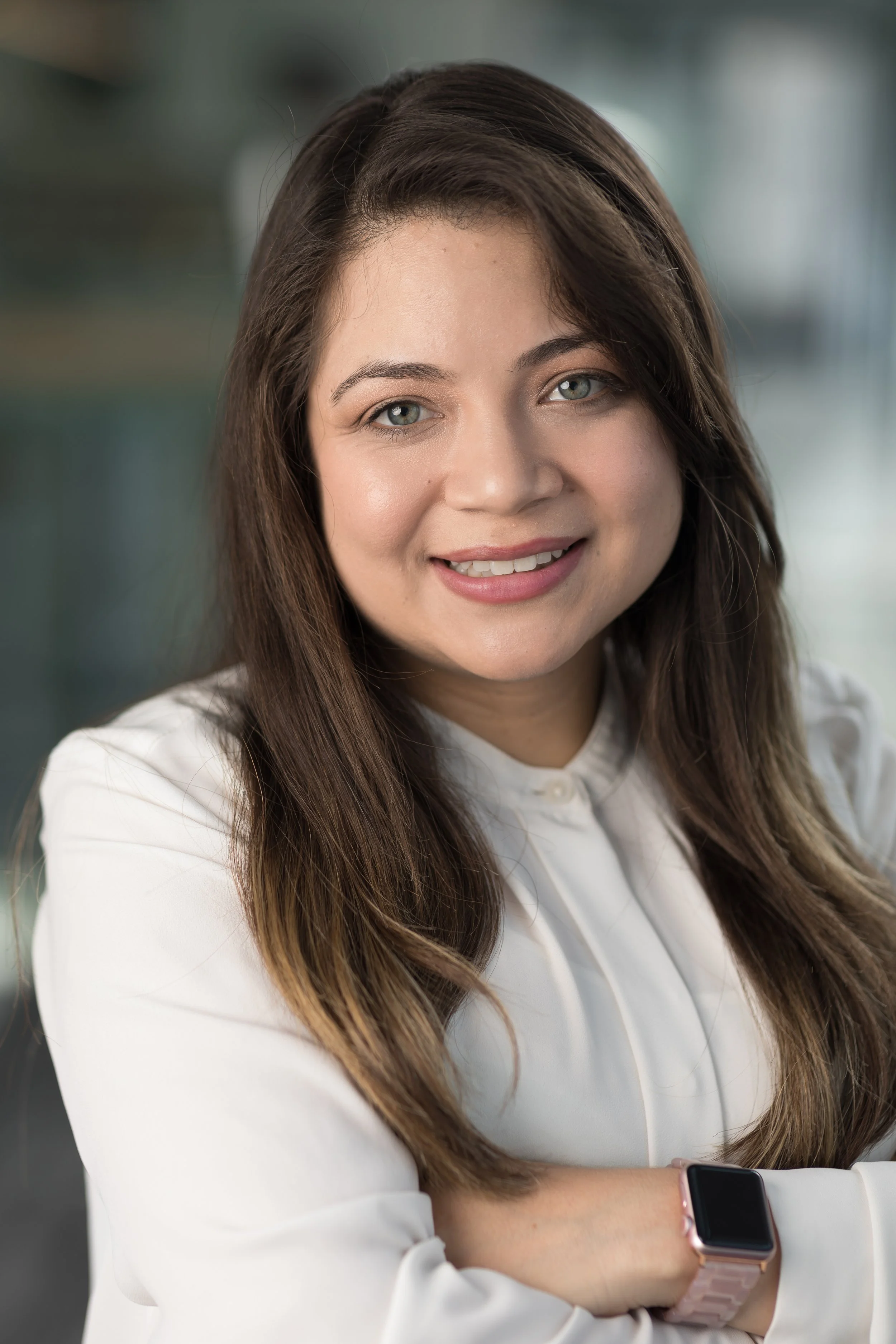 A woman with long brown hair wearing a white blouse, smiling, with arms crossed, and wearing a smartwatch on her left wrist in an indoor setting.