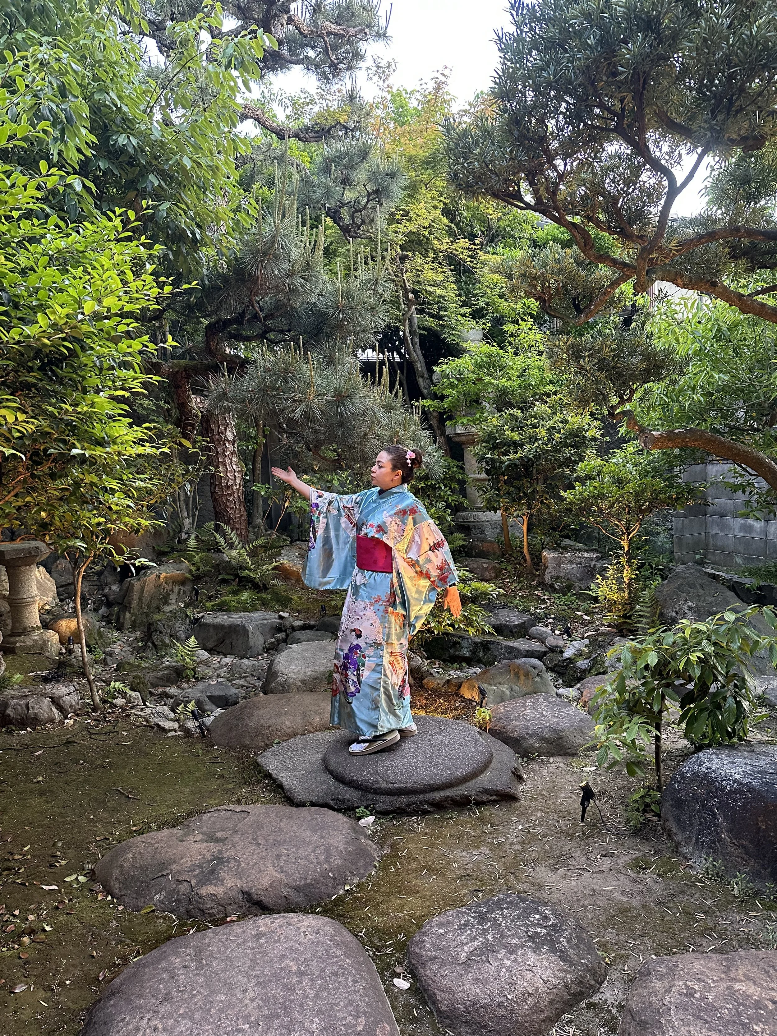 A woman dressed in a colorful kimono standing on large stones in a lush garden with various green trees and plants.