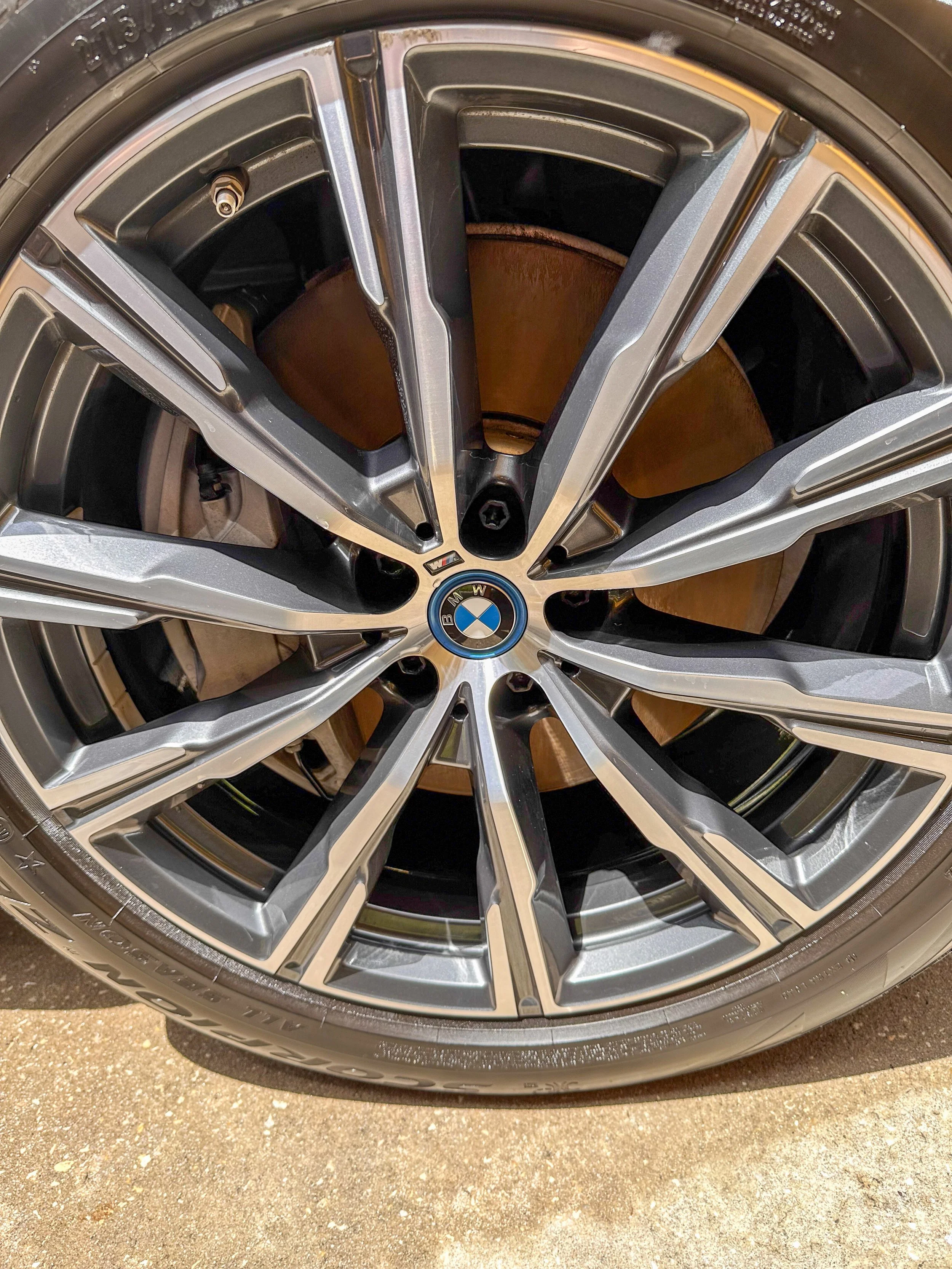 Close-up of a BMW car wheel with a black and silver alloy rim and a tire mounted on it, shown on a textured ground surface.