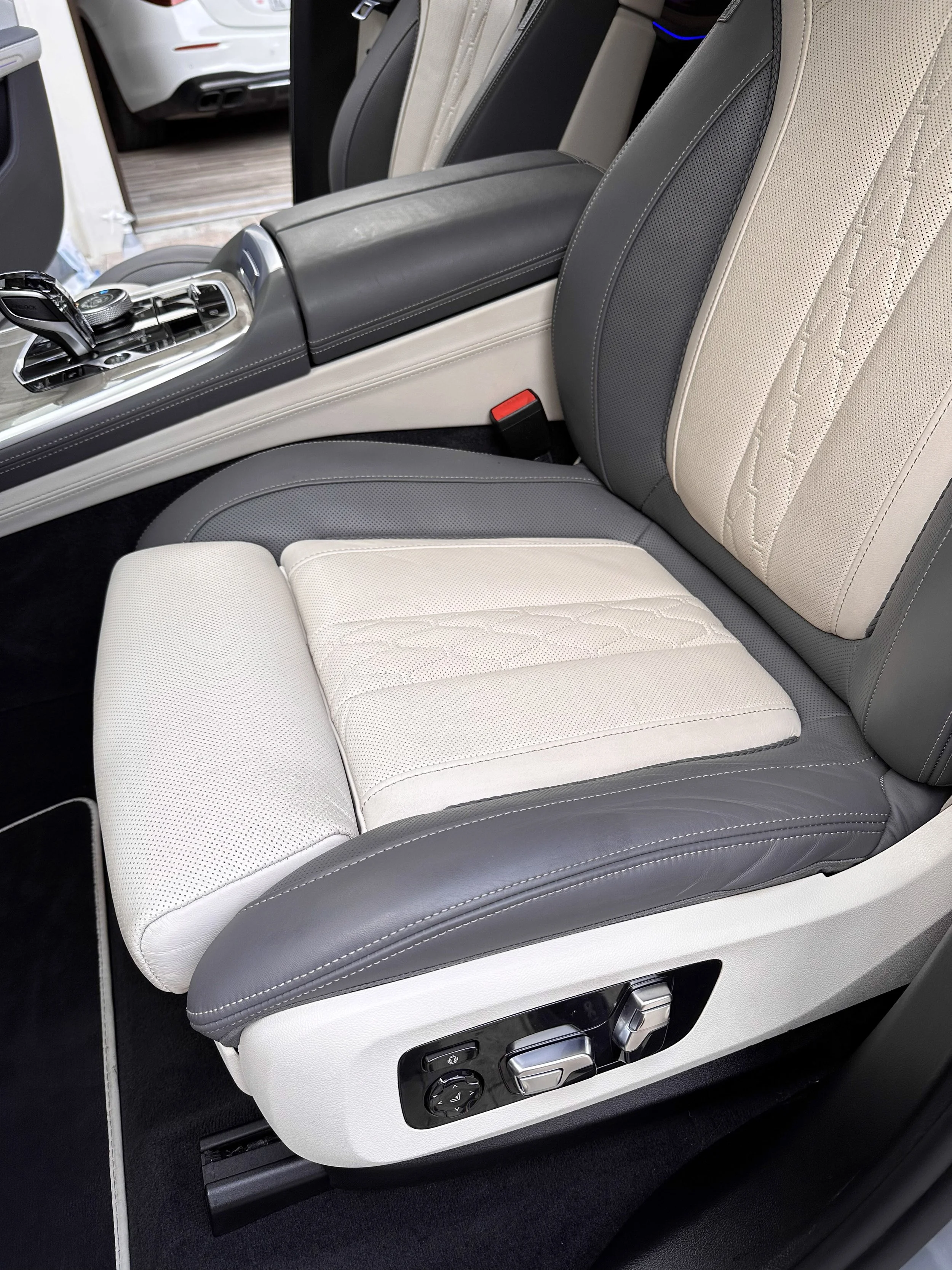 Close-up of a luxurious car seat with white and gray leather upholstery, electric seat adjustment controls on the side, and a center armrest with visible stitching.