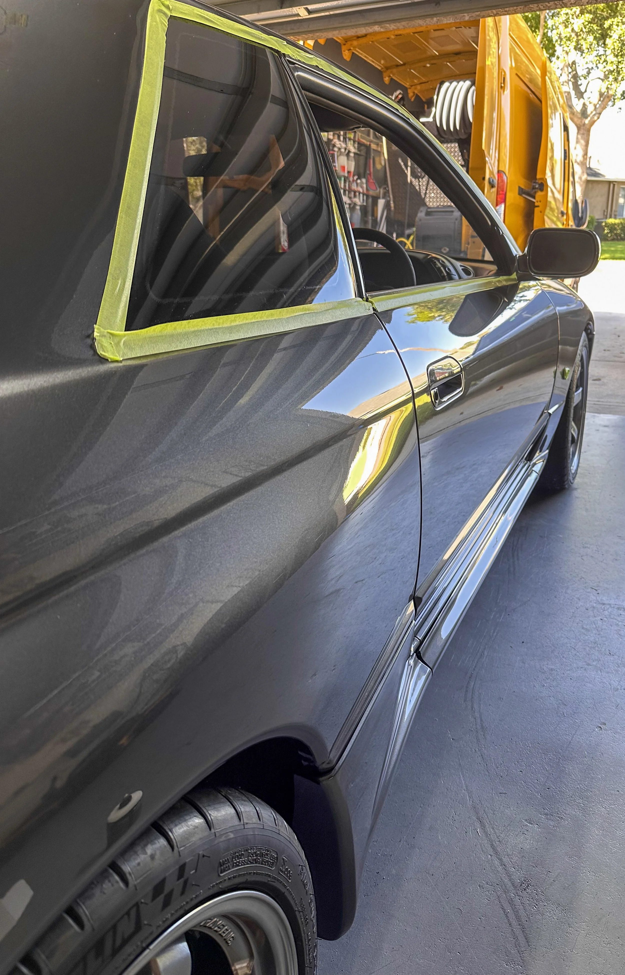 A black car with a yellow work vehicle in the background parked in a driveway, with trees and a house visible.