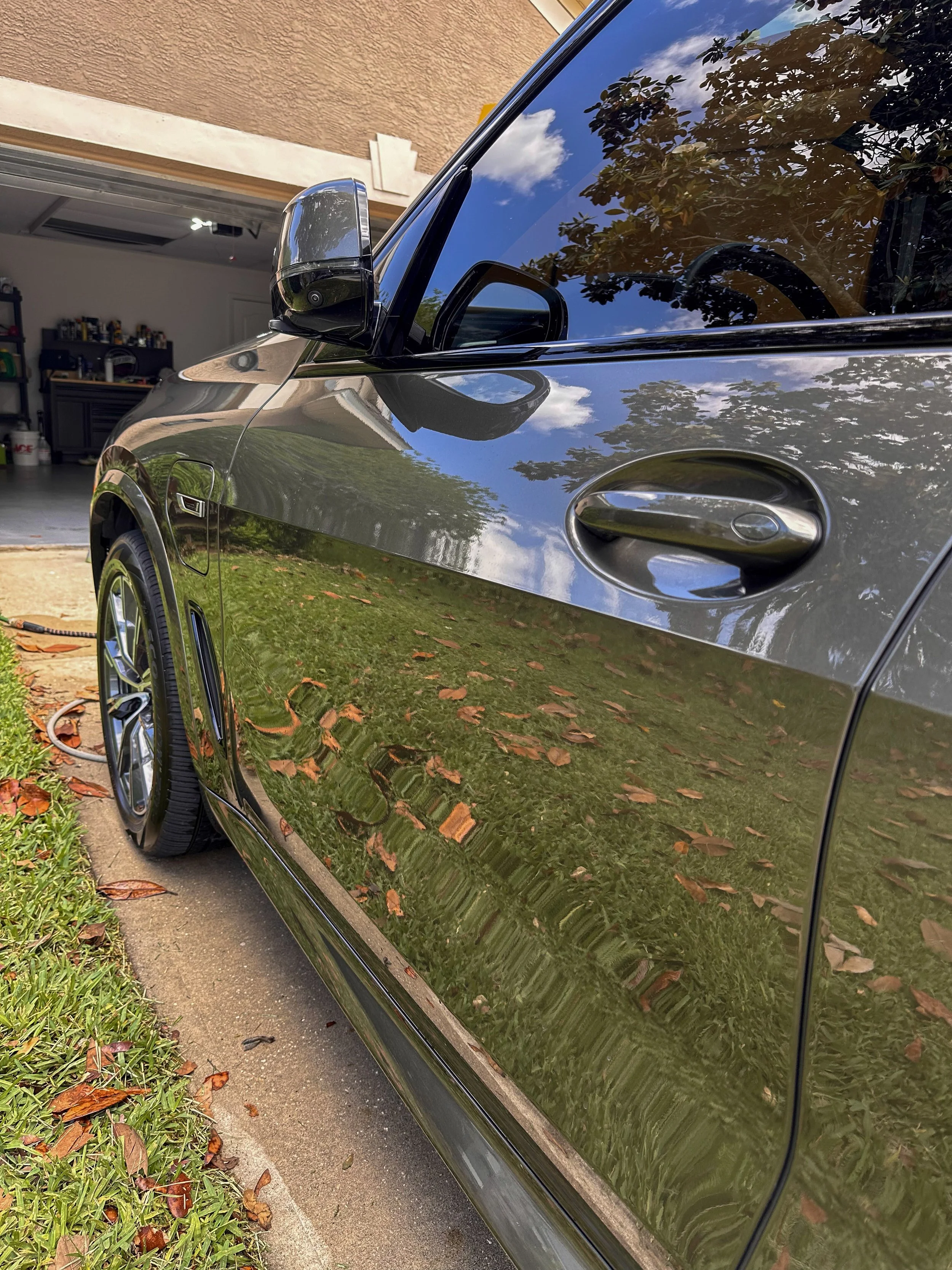 Reflection of trees and clouds on the shiny black side of a car parked on a grass and concrete driveway. In the background, a garage with an open door showing tools and supplies.