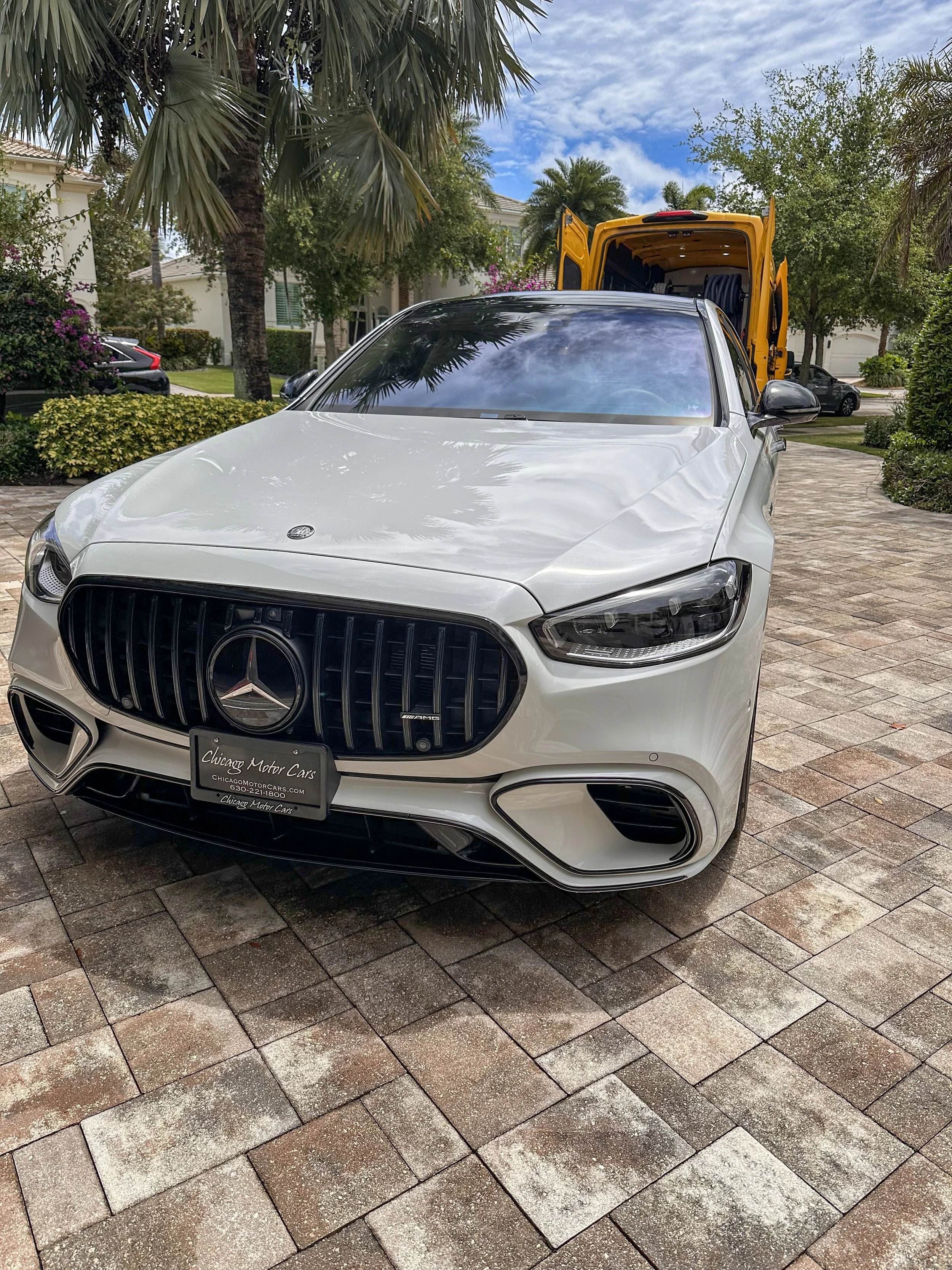 A white Mercedes-Benz car parked on a brick driveway in front of a lush suburban yard with palm trees and houses, with a yellow service vehicle behind it.