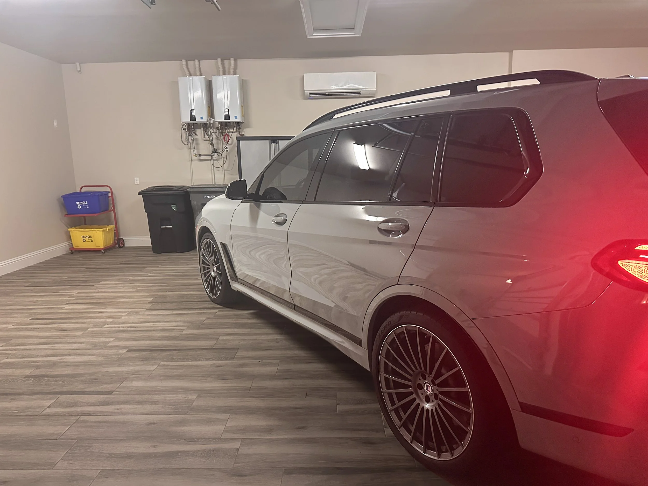 Garage interior with a parked silver luxury car, water heating units, recycling bins, and wood-look flooring.