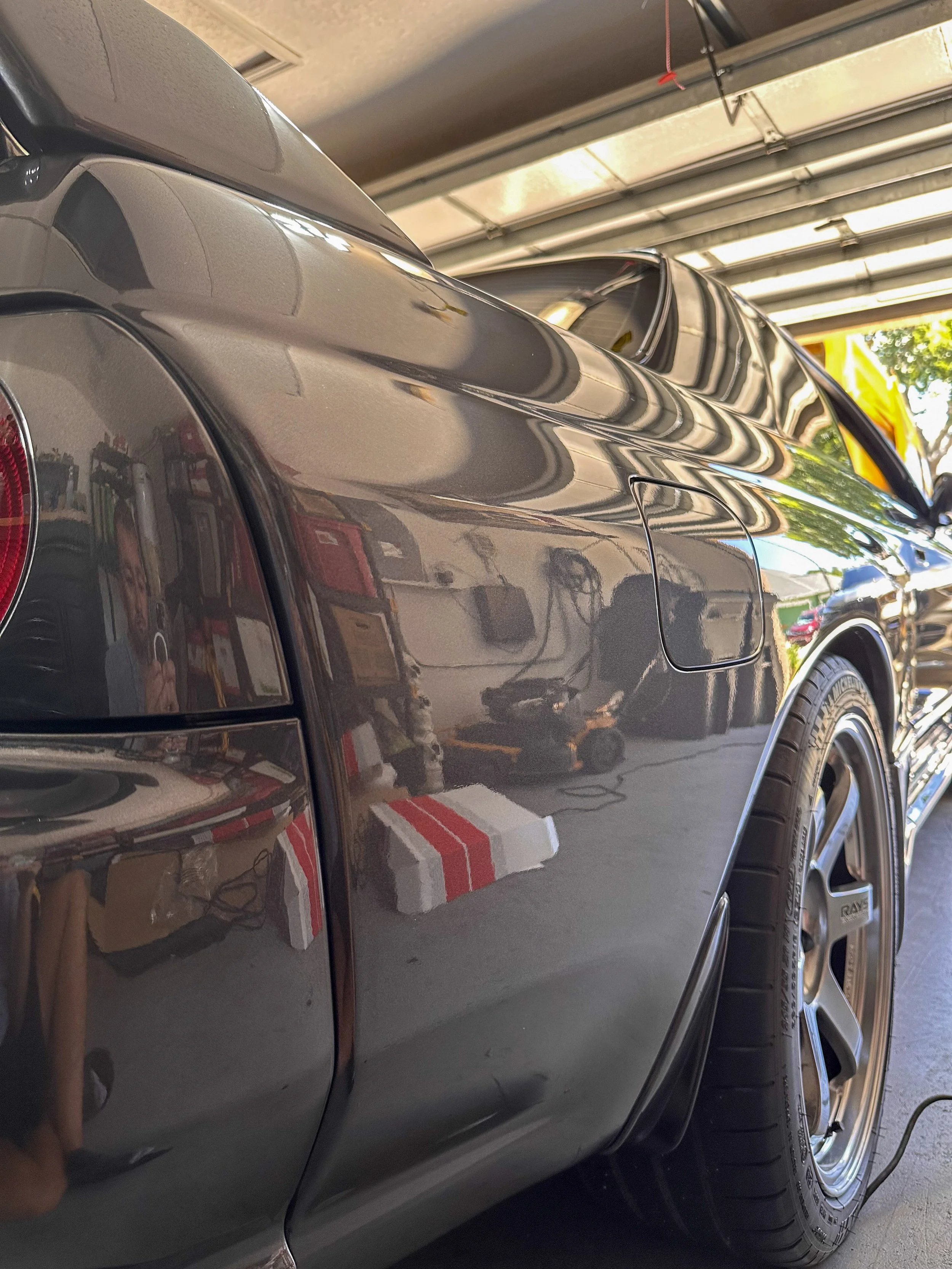Close-up of a shiny gray car in a garage, showing part of the rear quarter panel, wheel, and reflection of the garage interior.