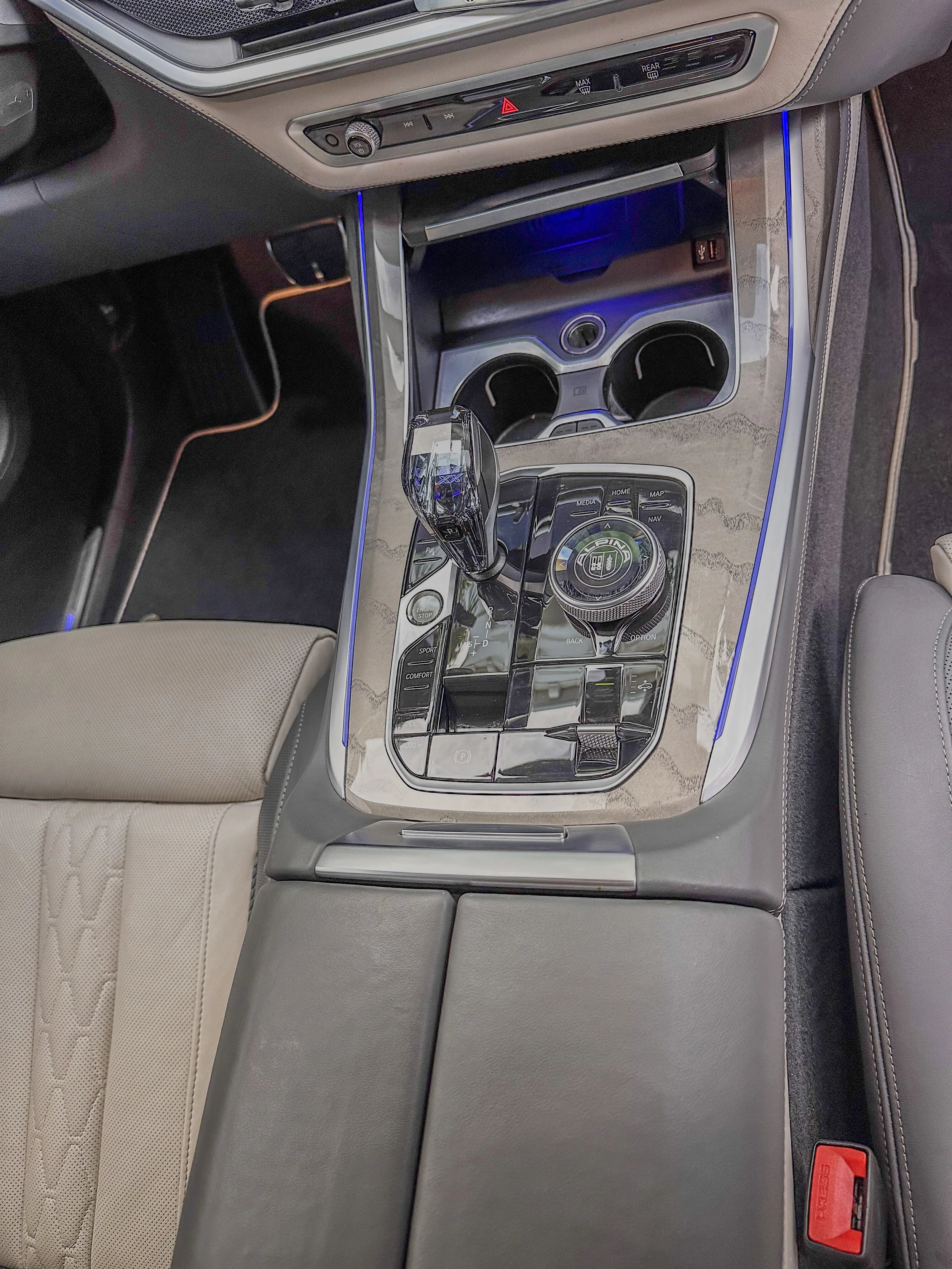 The interior of a luxury car showing the center console with a gear shifter, cup holders, and various control buttons, with beige and black leather upholstery and blue ambient lighting.