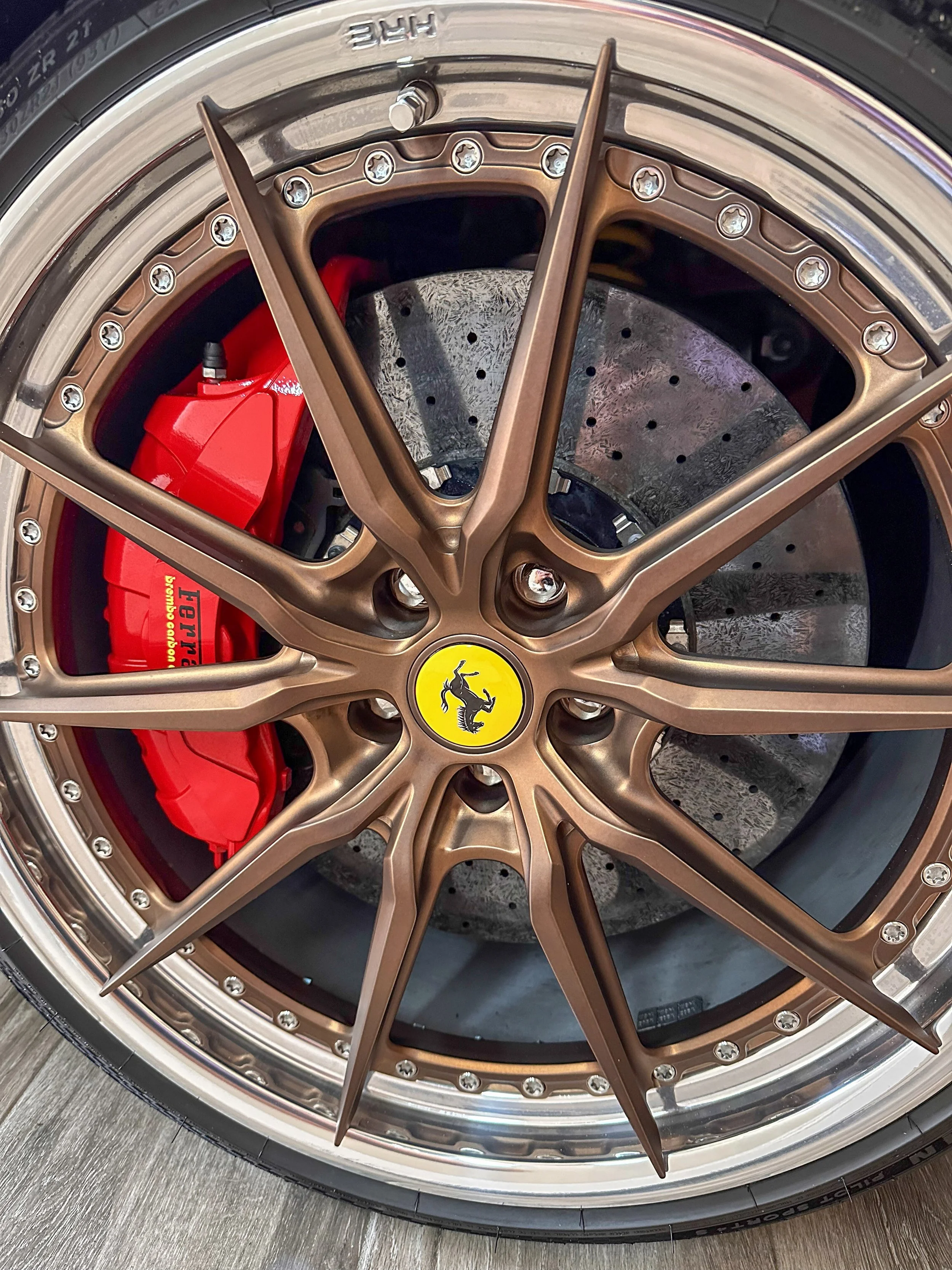 Close-up of a luxury car wheel featuring a bronze-colored rim with a Ferrari logo in the center, red brake caliper behind the rim, and a ventilated brake disc.