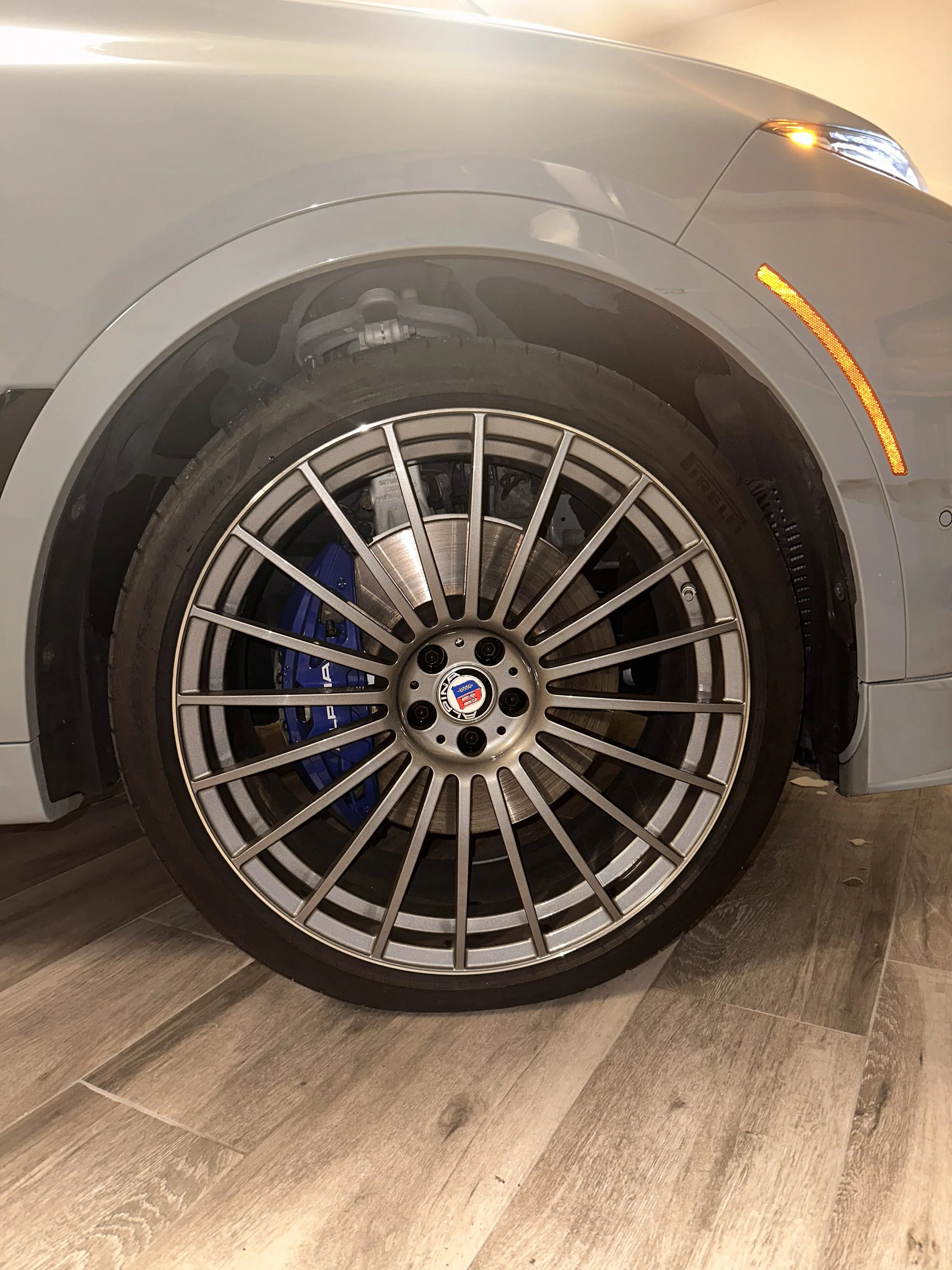 Close-up of the front wheel of a silver luxury car with a multi-spoke alloy rim, blue brake caliper, disc brake, and visible suspension components, parked on a wood floor.