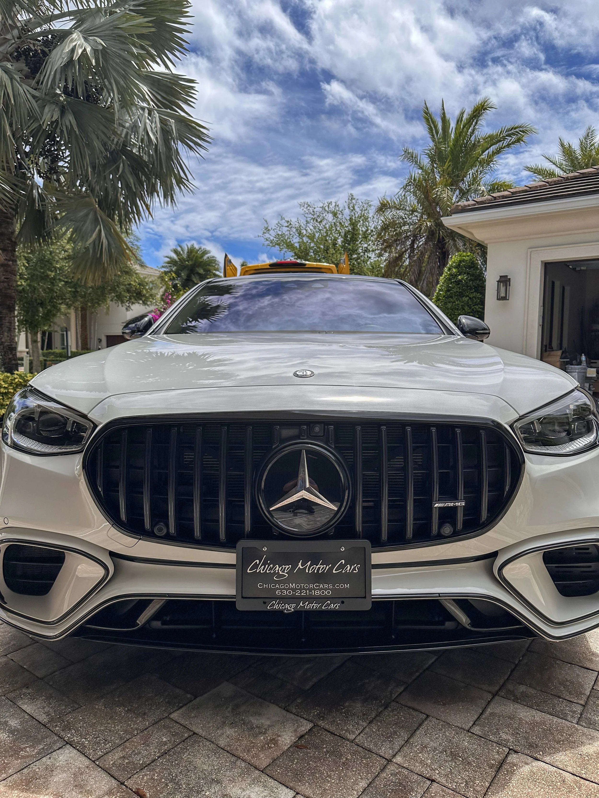 Front view of a white Mercedes-Benz car parked outside a house, with a palm tree and blue sky in the background, and a dealership plate reading 'Chicago Motor Cars'.