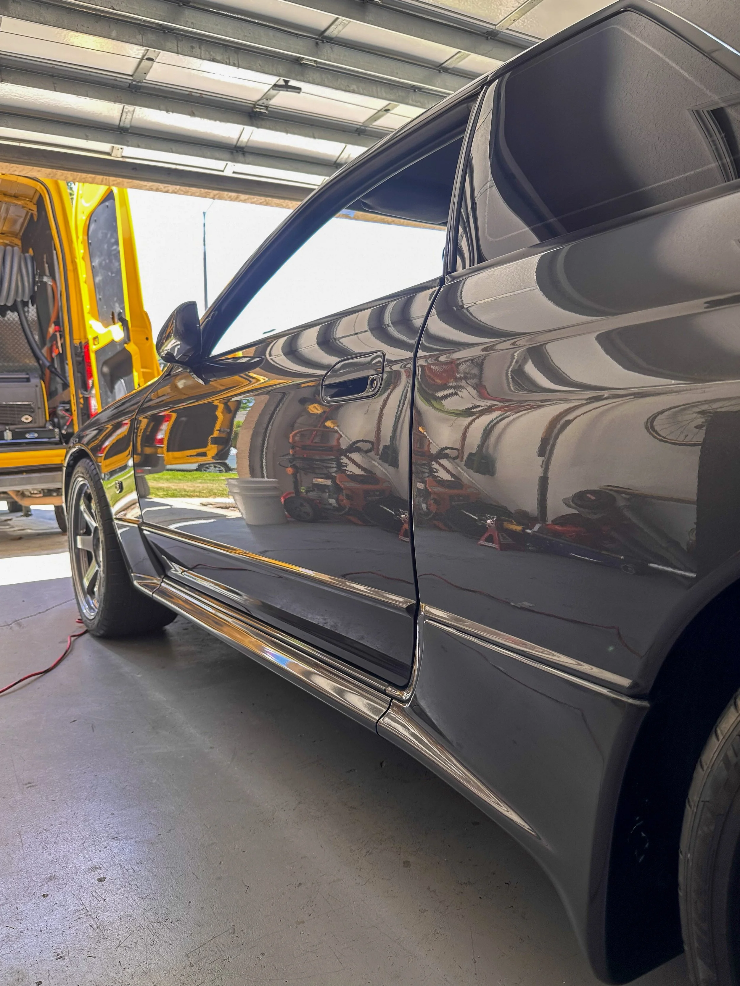 A black car with a glossy exterior parked in a garage, with reflections of the surroundings visible on the car's surface. To the left, there is a yellow service truck with open compartments containing tools and equipment.
