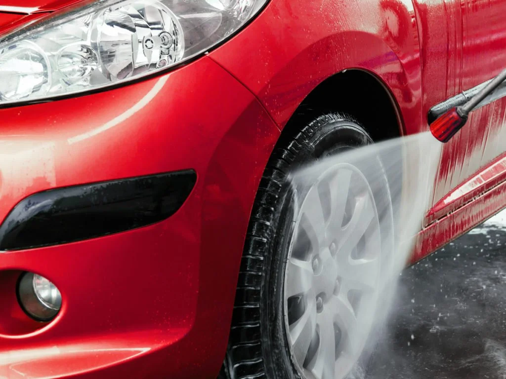 A red car is being washed with a pressure washer, water spraying on the front tire and side of the vehicle.