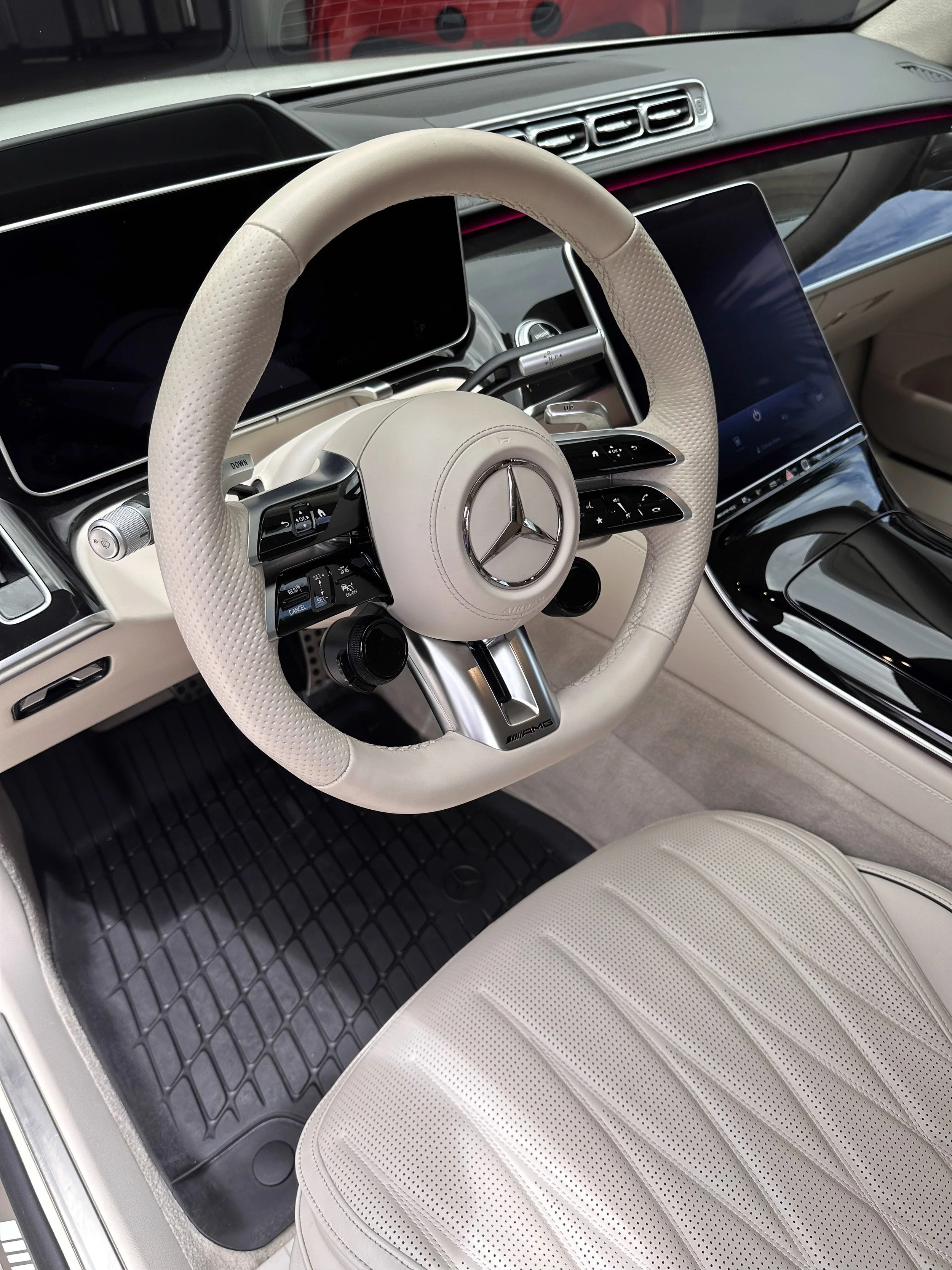 A luxury car's interior, focusing on the beige leather seat, white steering wheel with Mercedes-Benz logo, and large digital displays on the dashboard.