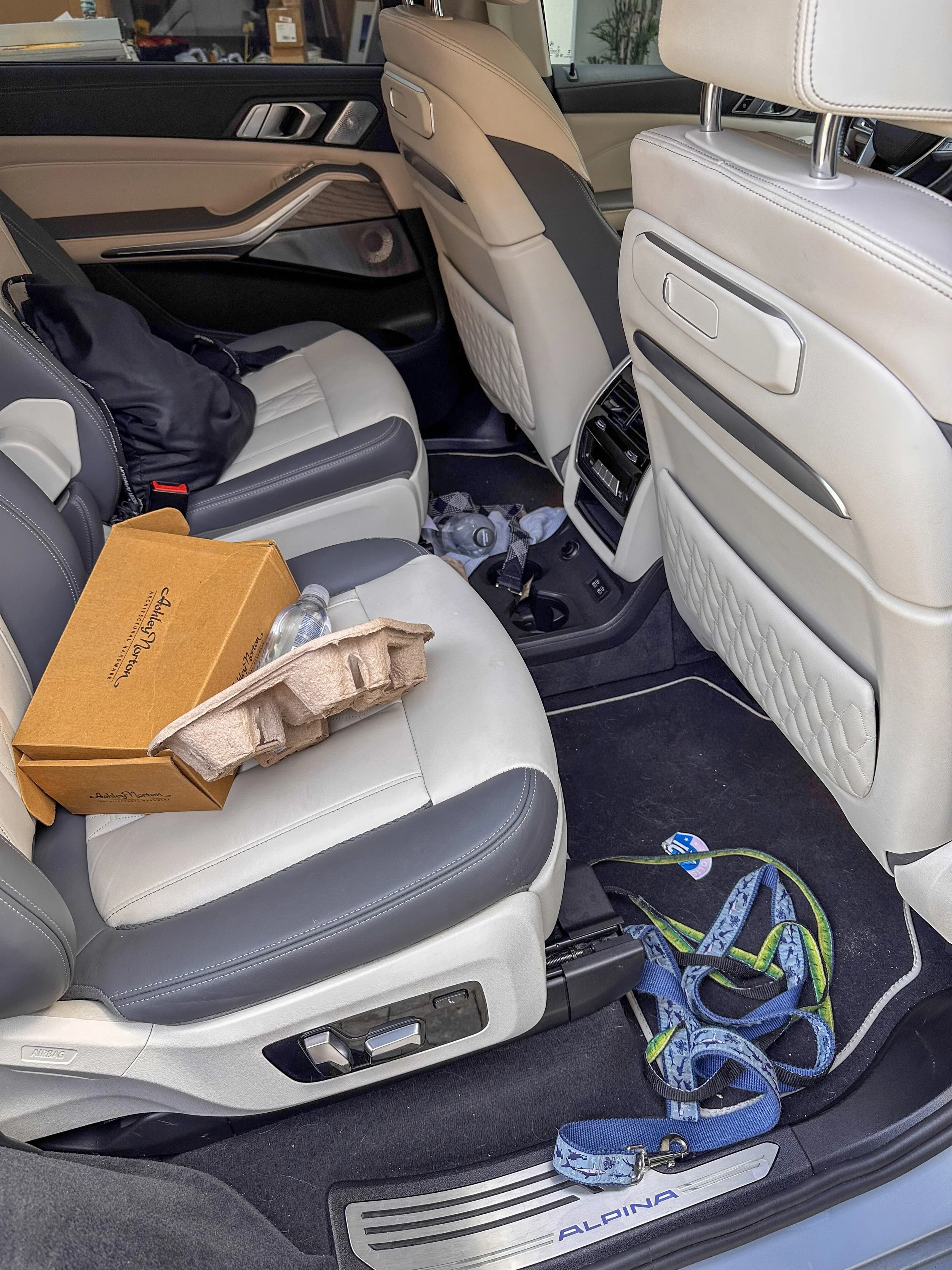 Inside a car showing the back of the front seats and the rear seat area, with some items such as a cardboard box, a water bottle, a bag, and a car leash on the seats and floor.