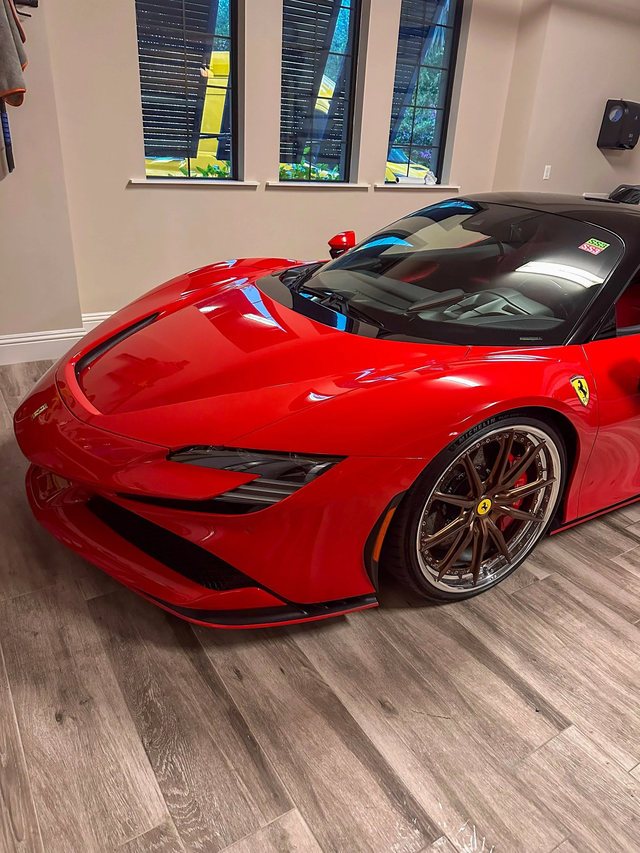 Red Ferrari sports car parked indoors with wooden flooring, with three large windows showing outside greenery and a yellow vehicle.