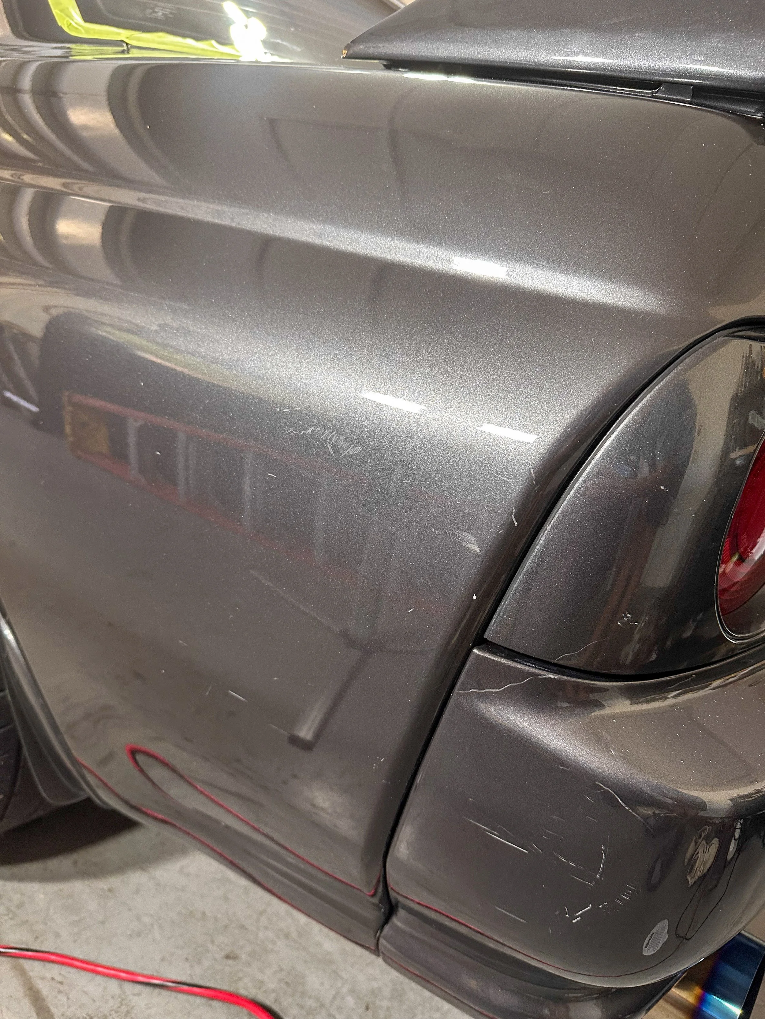 Close-up photo of the rear side of a dark gray or metallic gray car, showing the taillight, rear wheel with a crack, and some reflections on the paint.