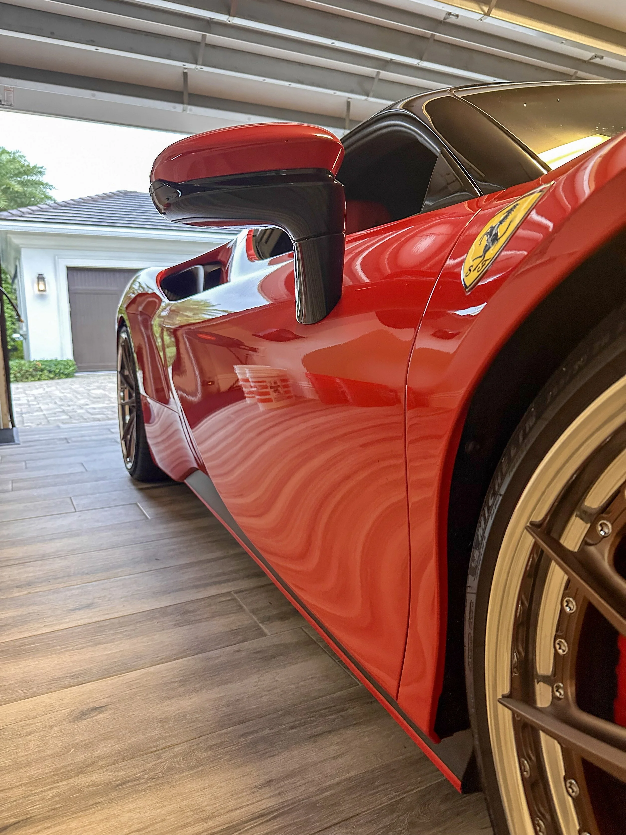 Close-up of a red Ferrari sports car with a yellow Ferrari badge and a gold-colored wheel, parked inside a garage with wooden flooring and an open garage door.