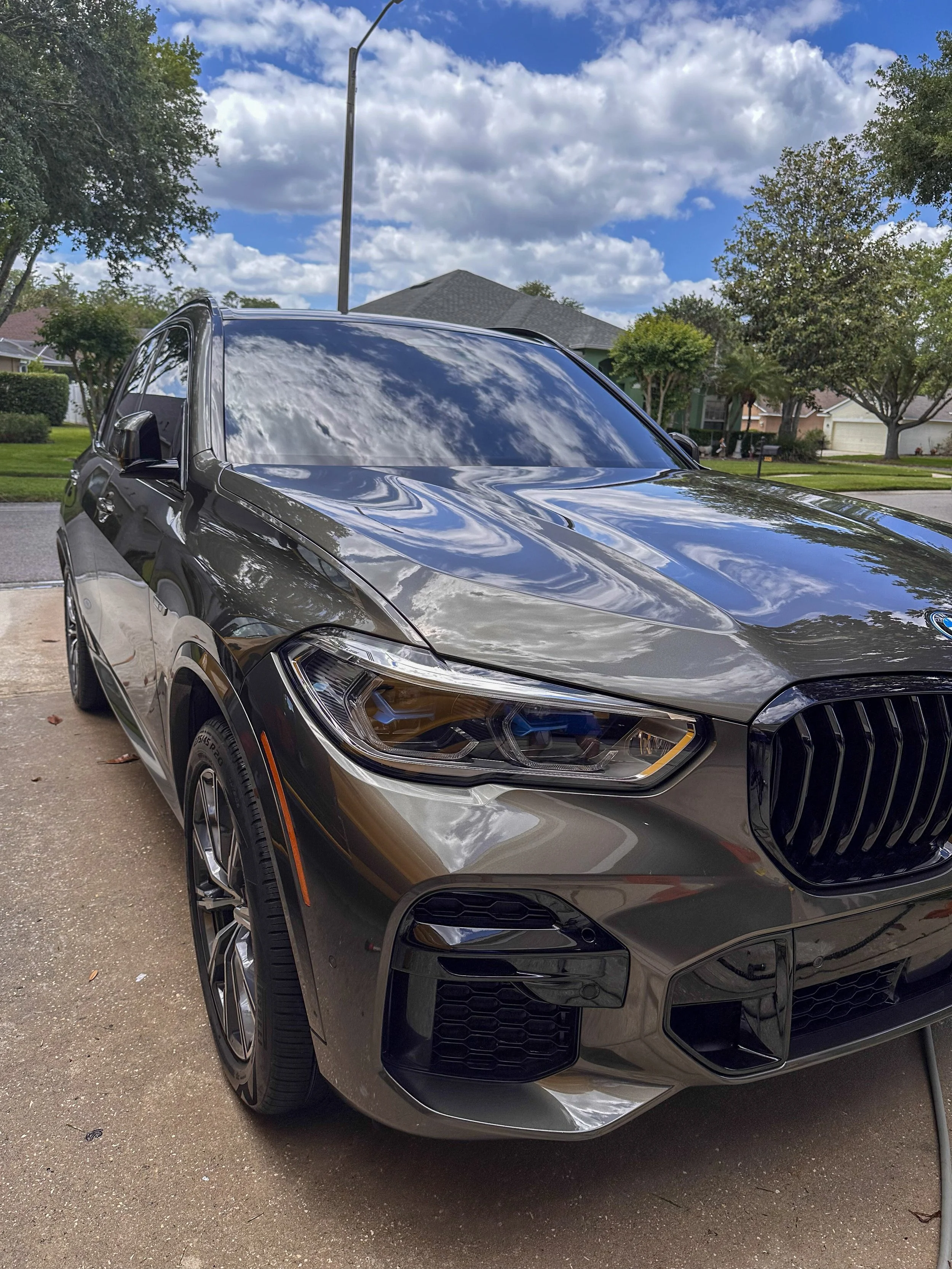 Close-up of a black modern car parked on a driveway in a suburban neighborhood with houses, trees, and a partly cloudy sky reflected on the car's windshield.