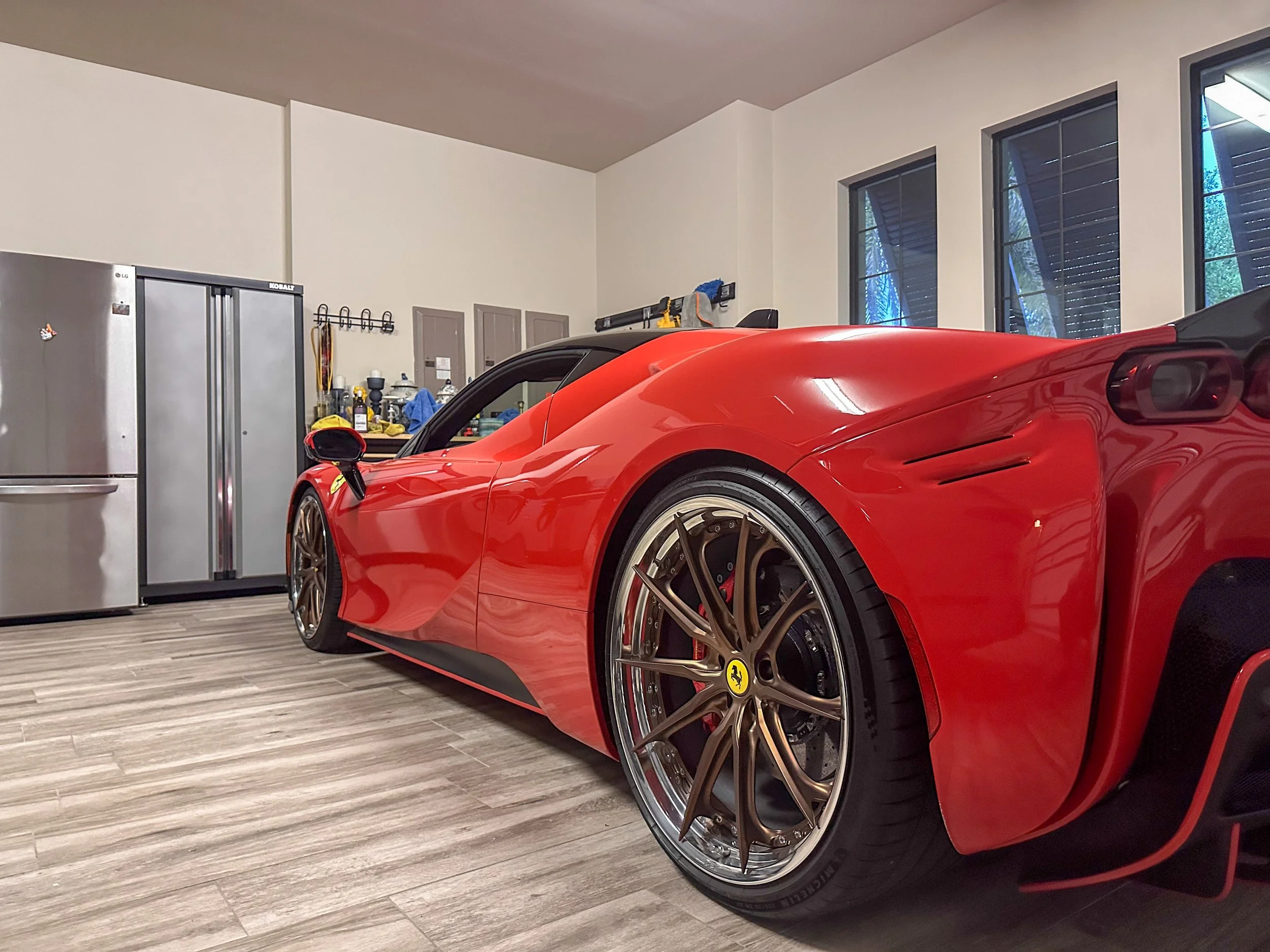 Close-up of a red luxury sports car, likely a Ferrari, parked in a modern indoor garage with large windows and a gray wooden floor. The car's front wheel and sleek body are prominently visible.