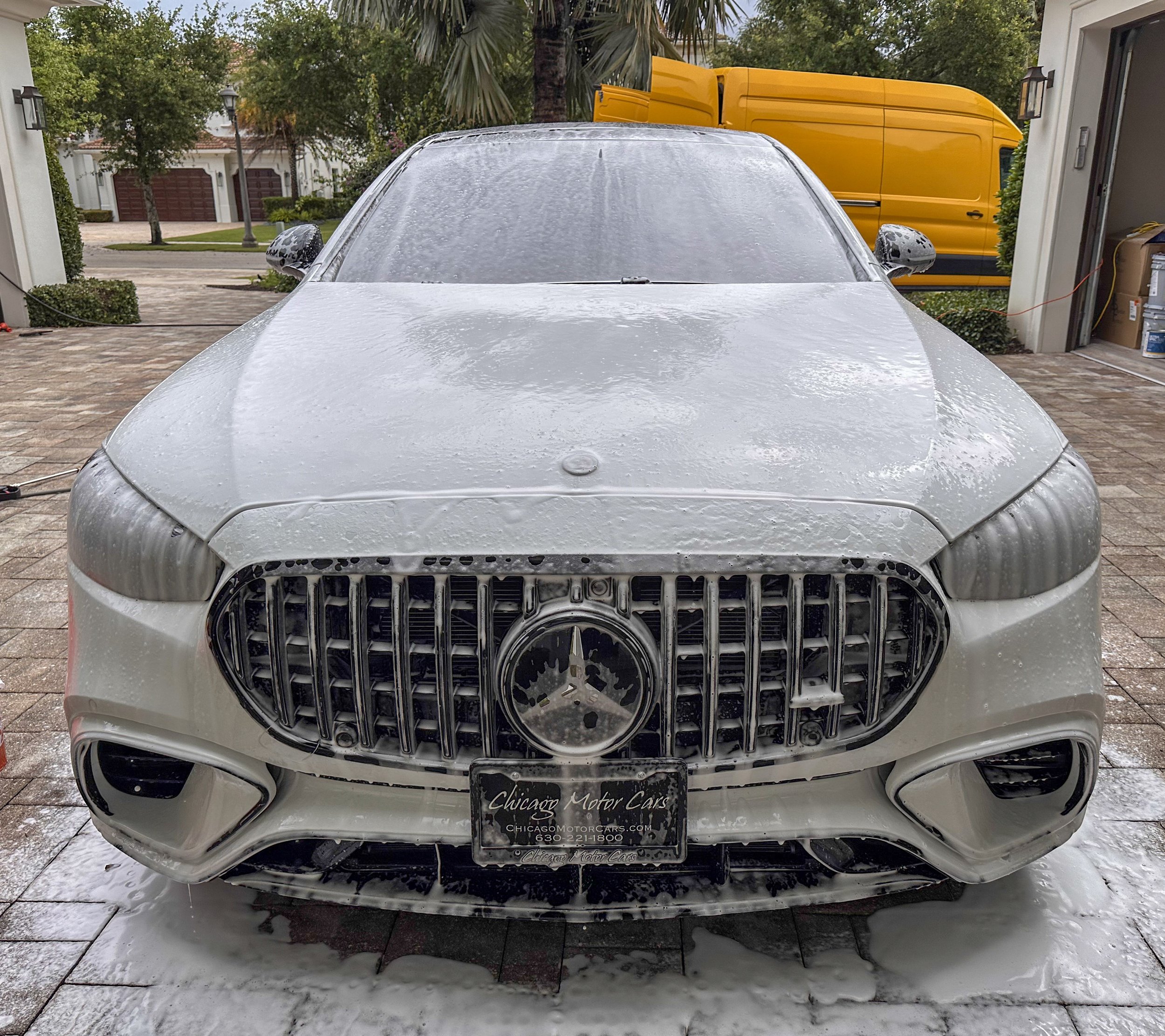 Front view of a white Mercedes-Benz car covered in soap suds during a car wash, with soap foam on the windshield and hood, in a driveway with trees and a house in the background.