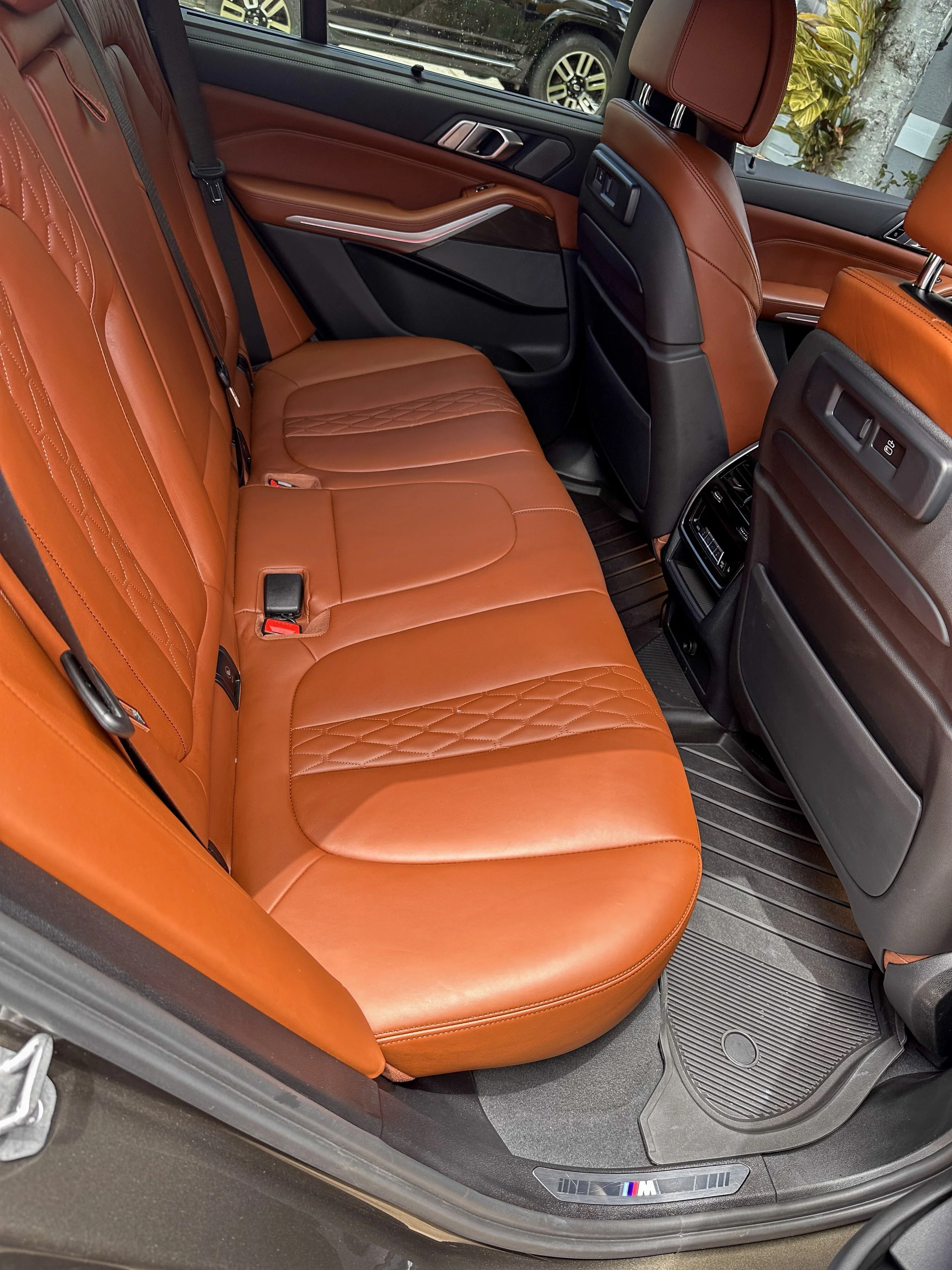 The back seat area of a luxury car with tan leather upholstery, detailed stitching, and a black interior design. The car's window shows a glimpse of a black car and green foliage outside, with the BMW M badge visible on the door sill.