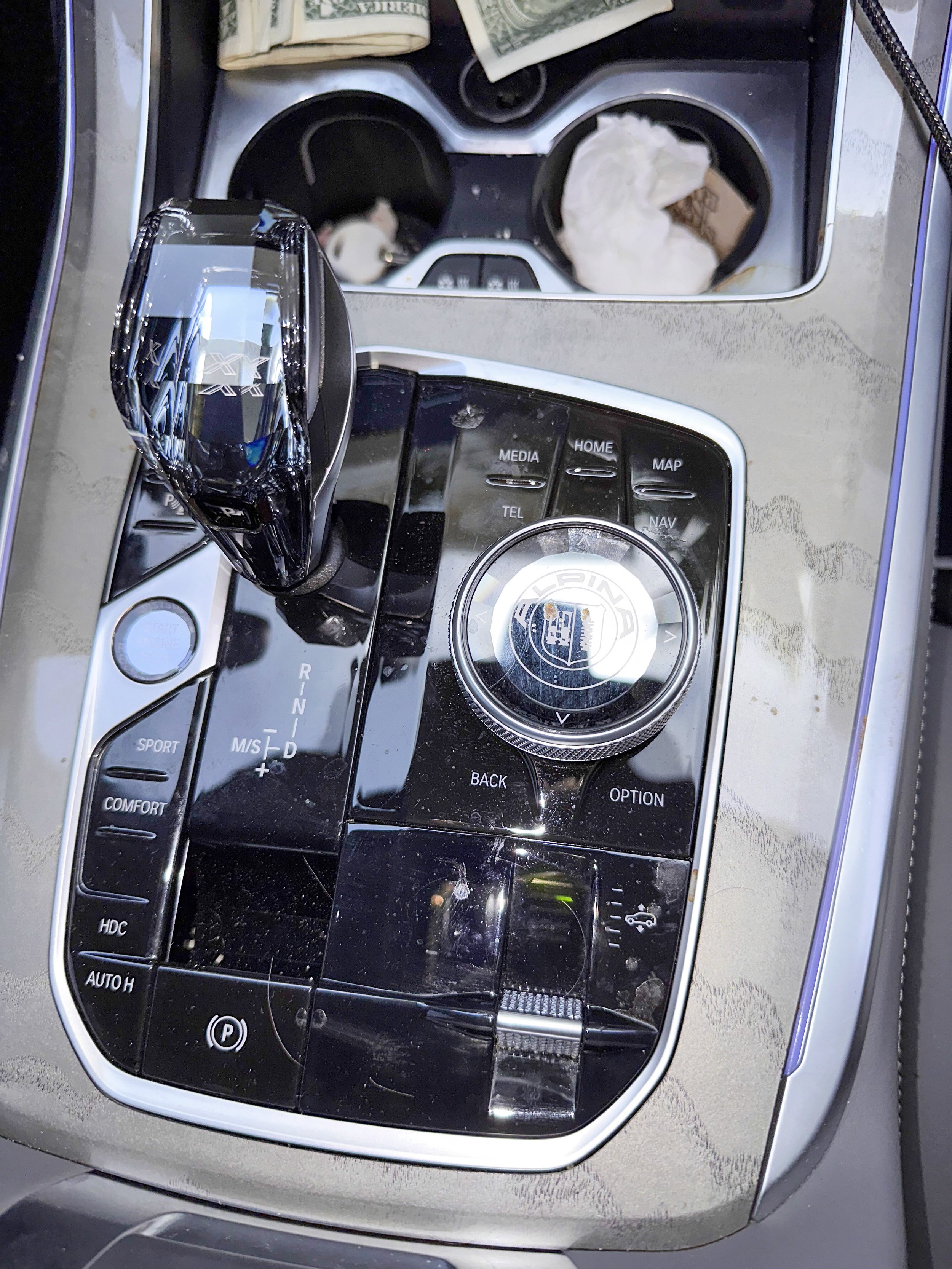 Close-up of a car's center console with gear shift, various control buttons, a rotary knob, and an open cup holder containing paper and McDonald's napkins. A blurred receipt or paper is seen in the top part of the image.