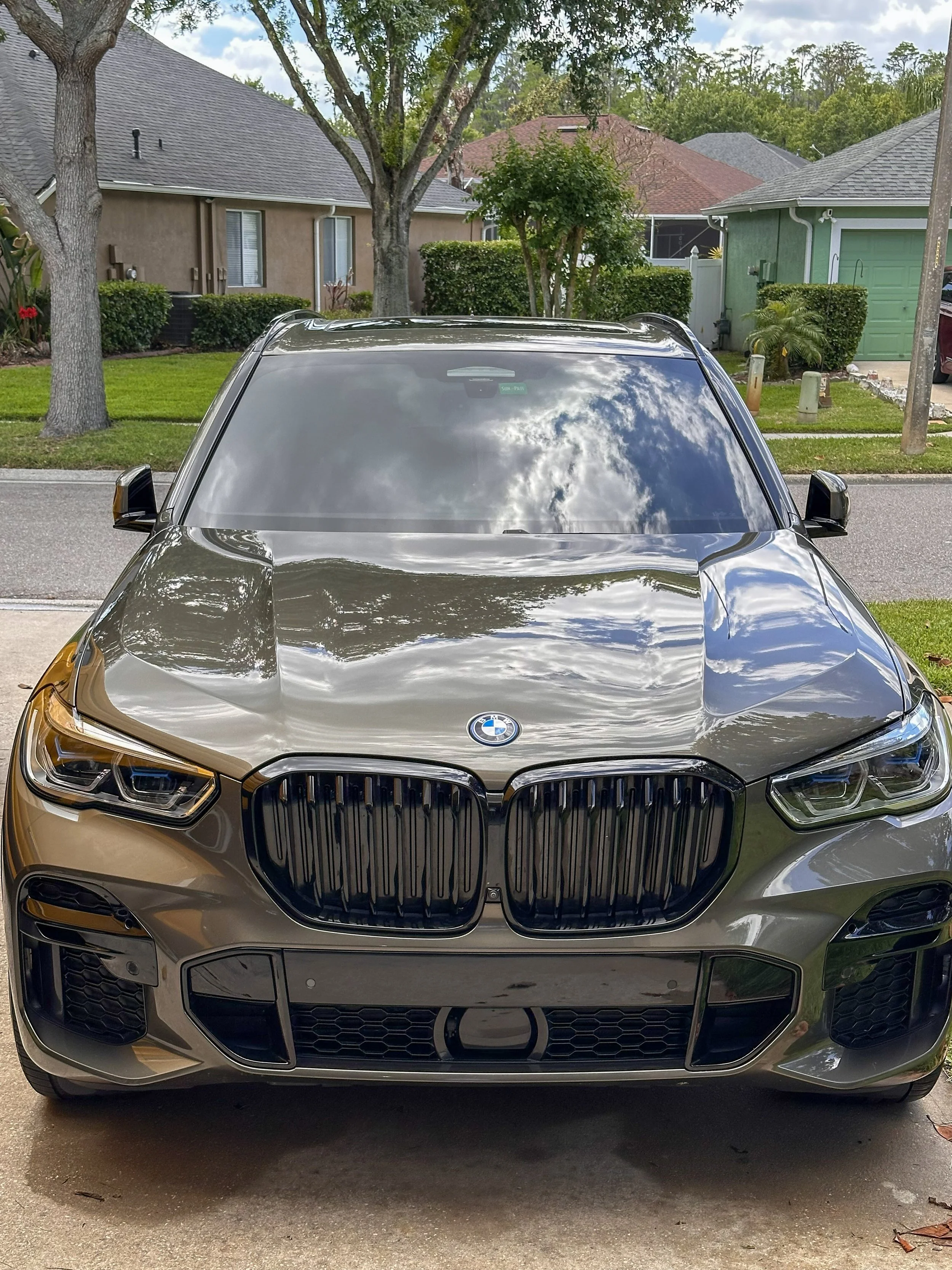 Front view of a shiny black BMW car parked on a residential driveway, with houses, trees, and a cloudy sky reflected on its windshield.