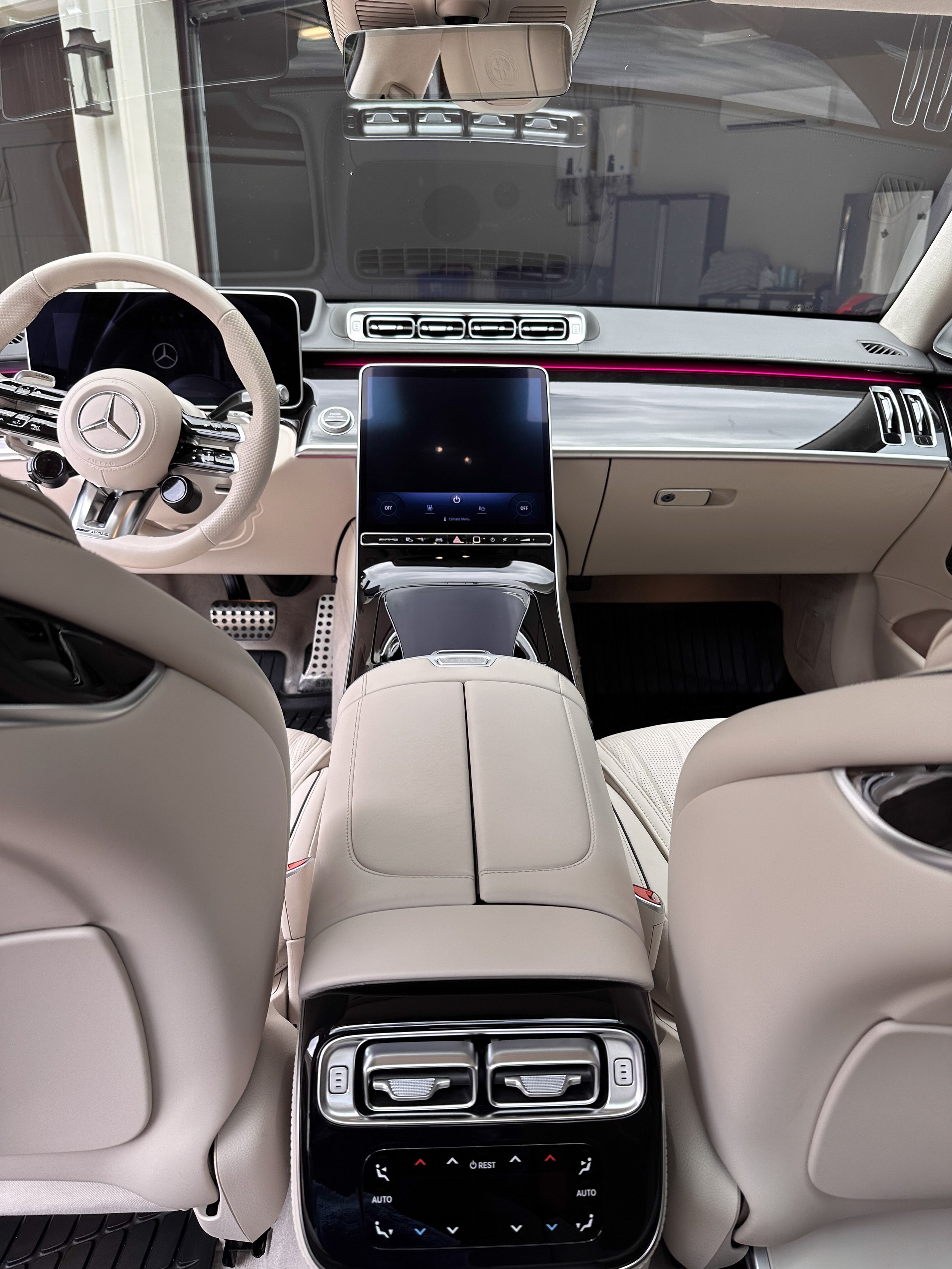 The inside of a luxury Mercedes-Benz vehicle, showing the beige leather seats, a central touchscreen display, rear climate control vents, and a rear center console with touch controls and vents.