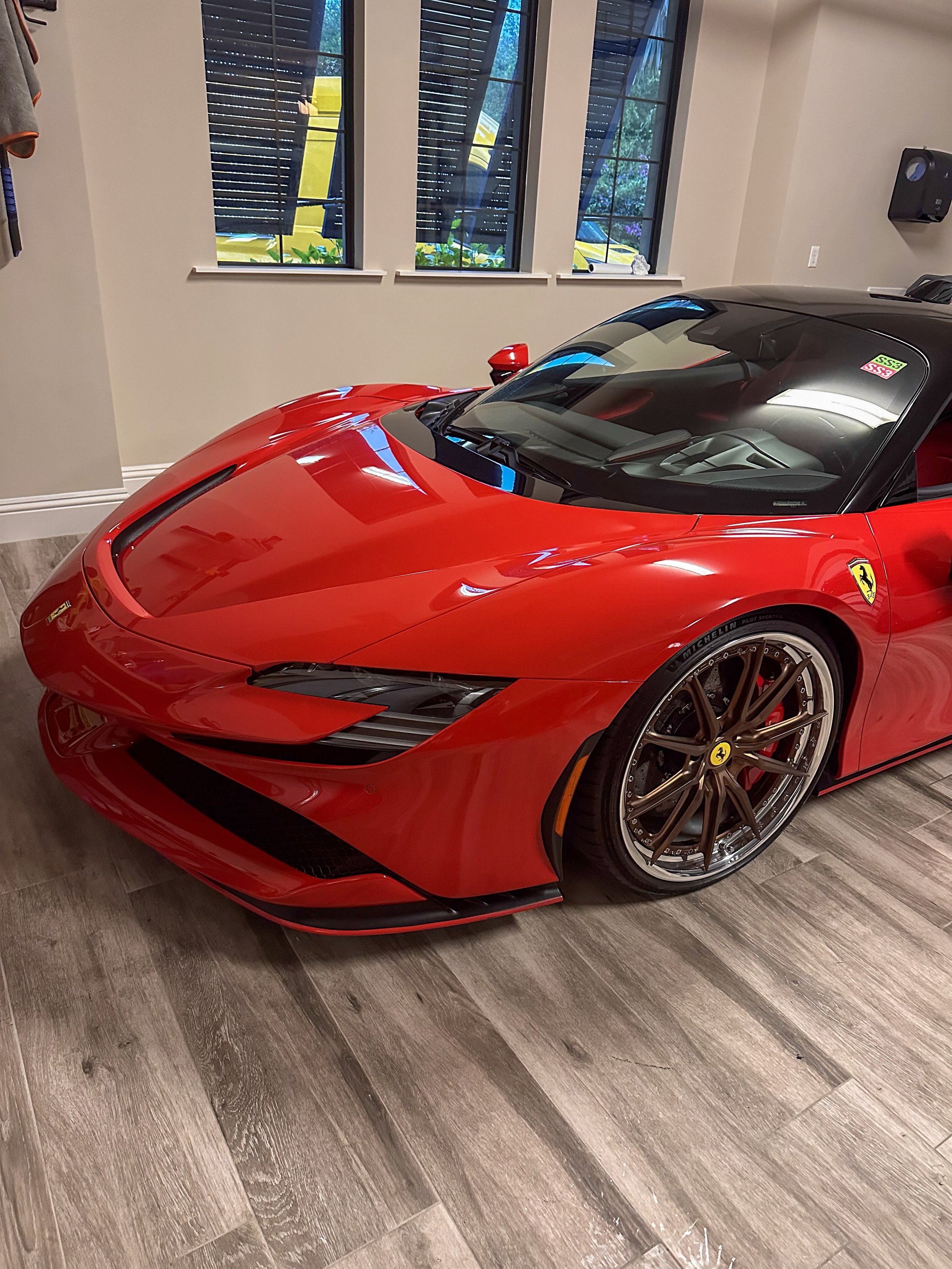 Red Ferrari sports car parked indoors on a wooden floor, with a black roof and tires bearing Michelin Pilot Sport 4S. The car has Ferrari logos on the sides and appears to be a modern model.