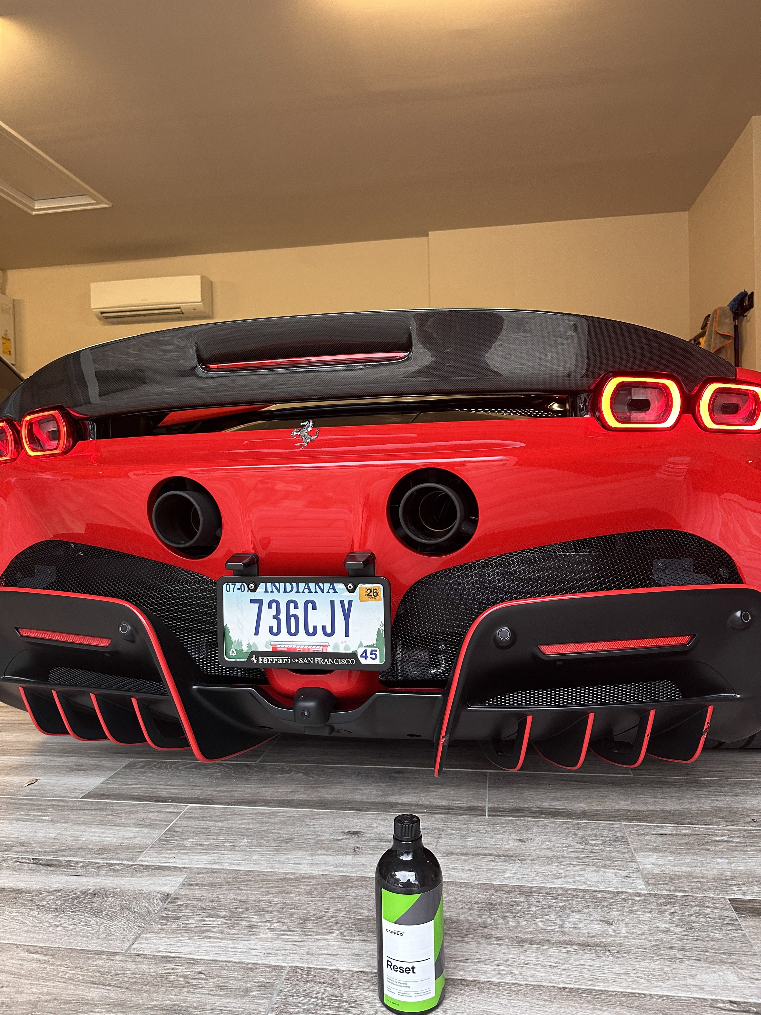 Rear view of a red Ferrari sports car with a black spoiler and rear diffuser, parked indoors against a beige wall, with an Indiana license plate reading 736C JY, and a black bottle with a green label on the wooden floor in front.