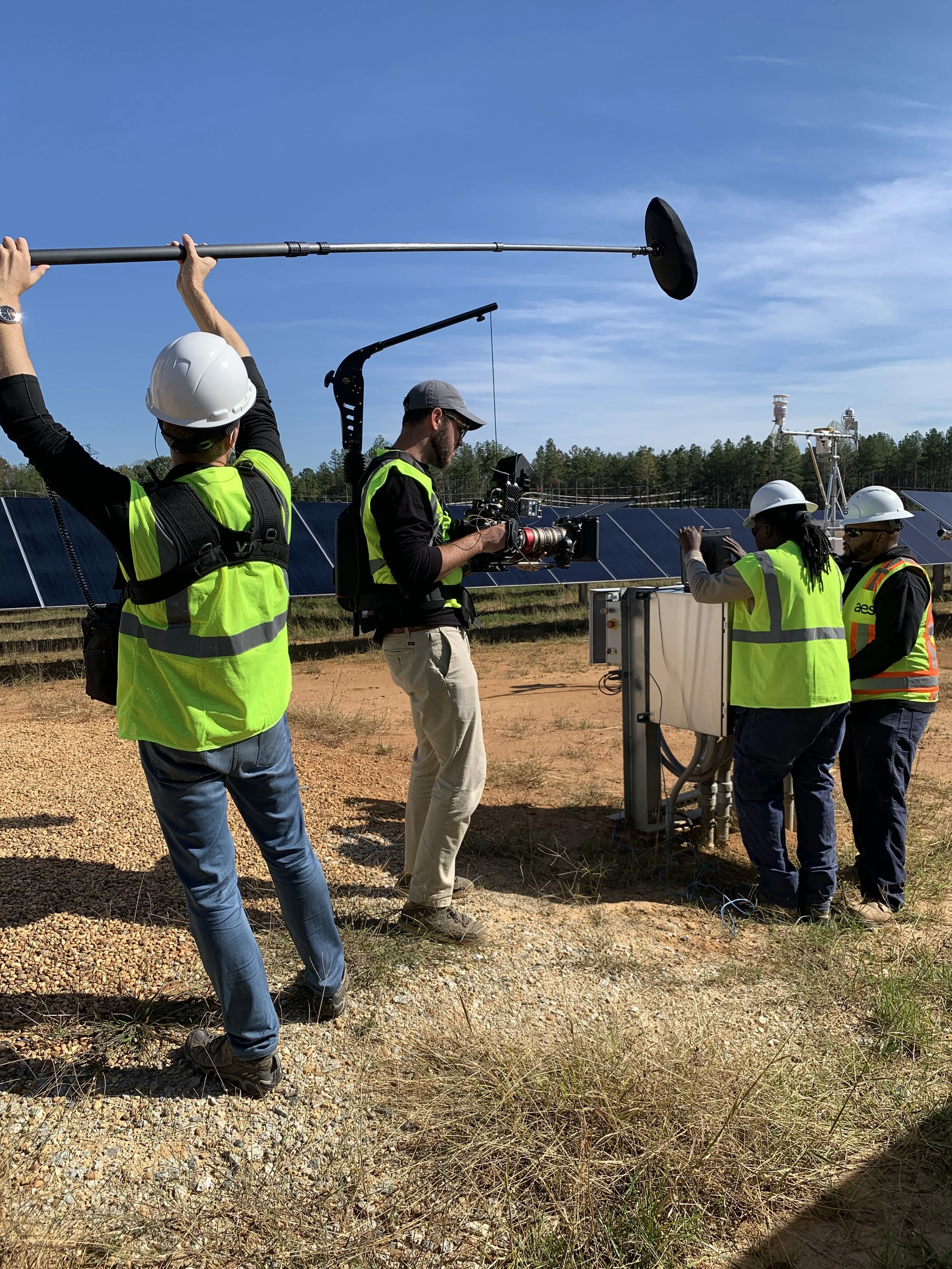 A group of people wearing safety vests and hard hats working outdoors near solar panels, with one person operating a camera on a crane, and others working with equipment.